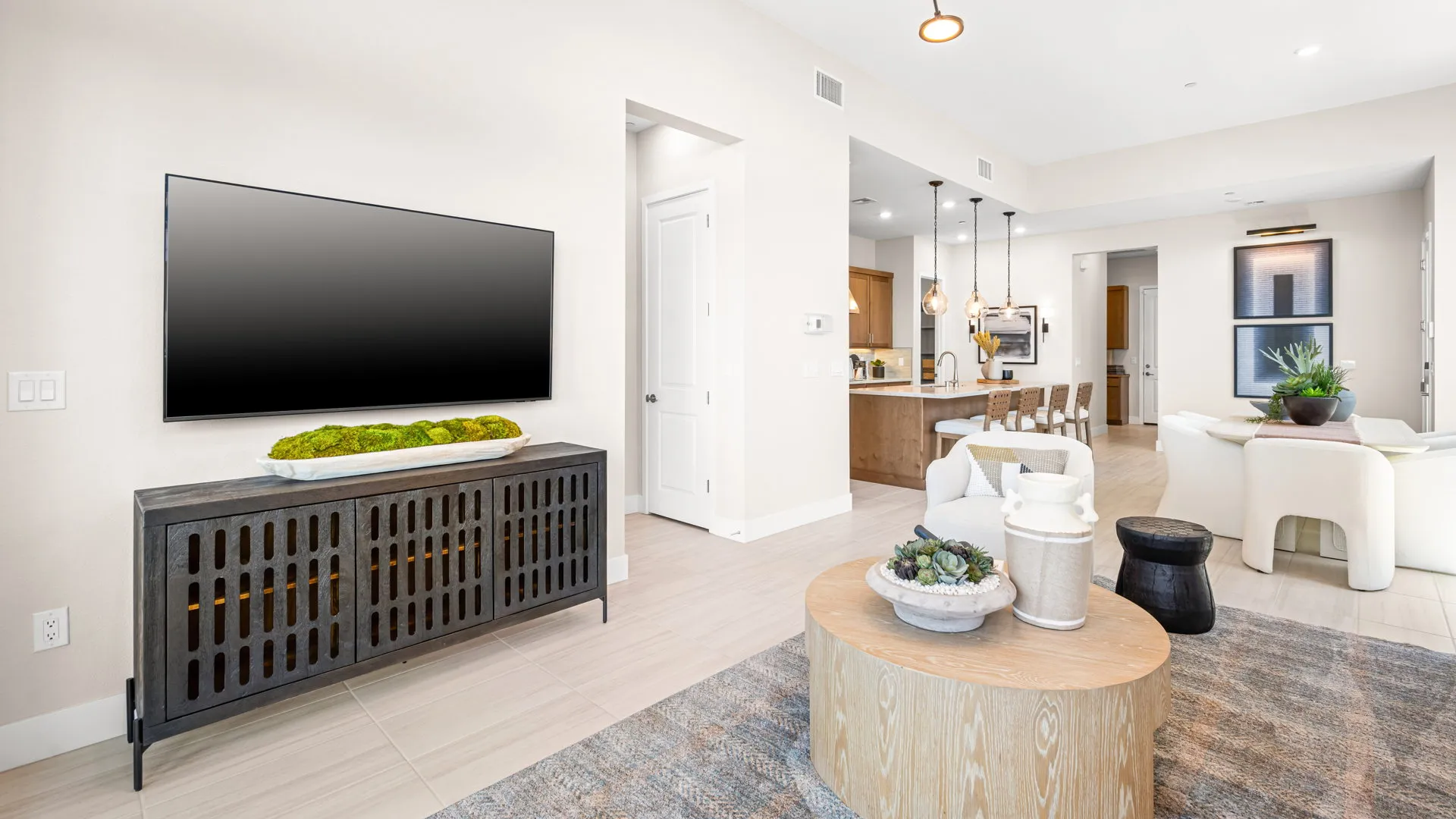 Living Room of Tiki Model at Esplanade at Red Rock by Taylor Morrison (2) Modern living room with a wall-mounted TV, console table, round wooden coffee table, white chairs, decorative plants, and an open kitchen with pendant lights in the background.