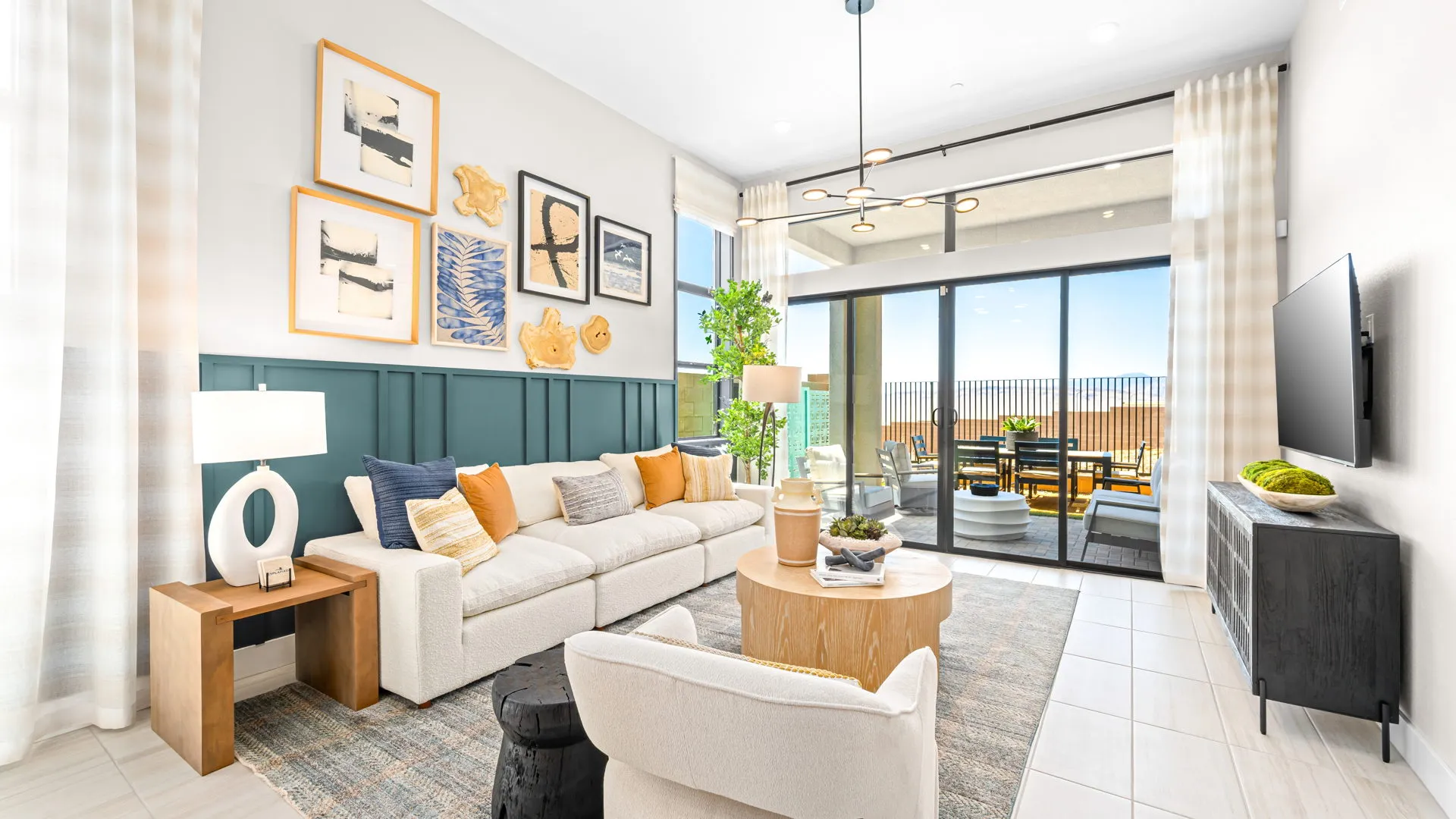 Living Room of Tiki Model at Esplanade at Red Rock by Taylor Morrison Bright, modern living room with white sofa, accent pillows, wall art, coffee table, armchair, TV, and large windows leading to a patio with outdoor seating and a scenic view. Neutral tones and green accents throughout.