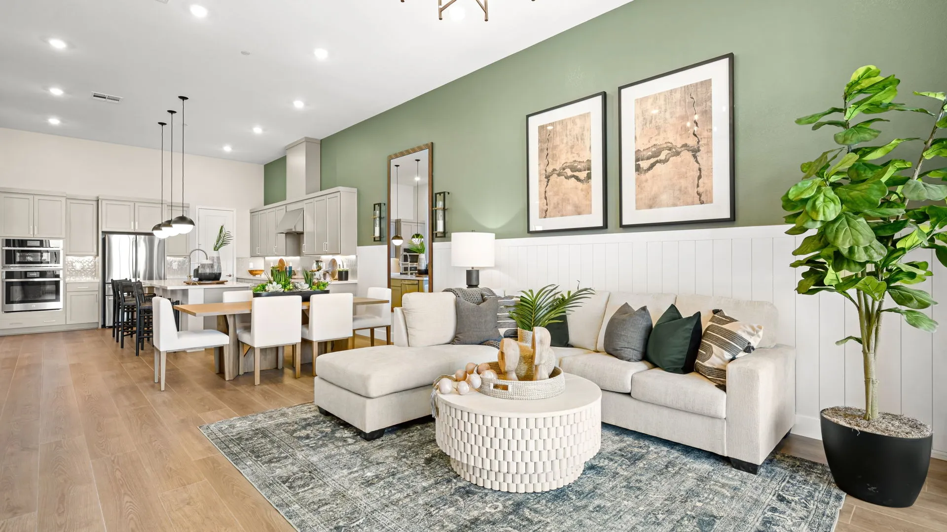 Living Room of Zinnia Model at Esplanade at Red Rock by Taylor Morrison (1) Open-concept living room and kitchen with green accent wall, beige sectional sofa, round textured coffee table, modern decor, dining area with white chairs, and wood flooring under bright lighting.