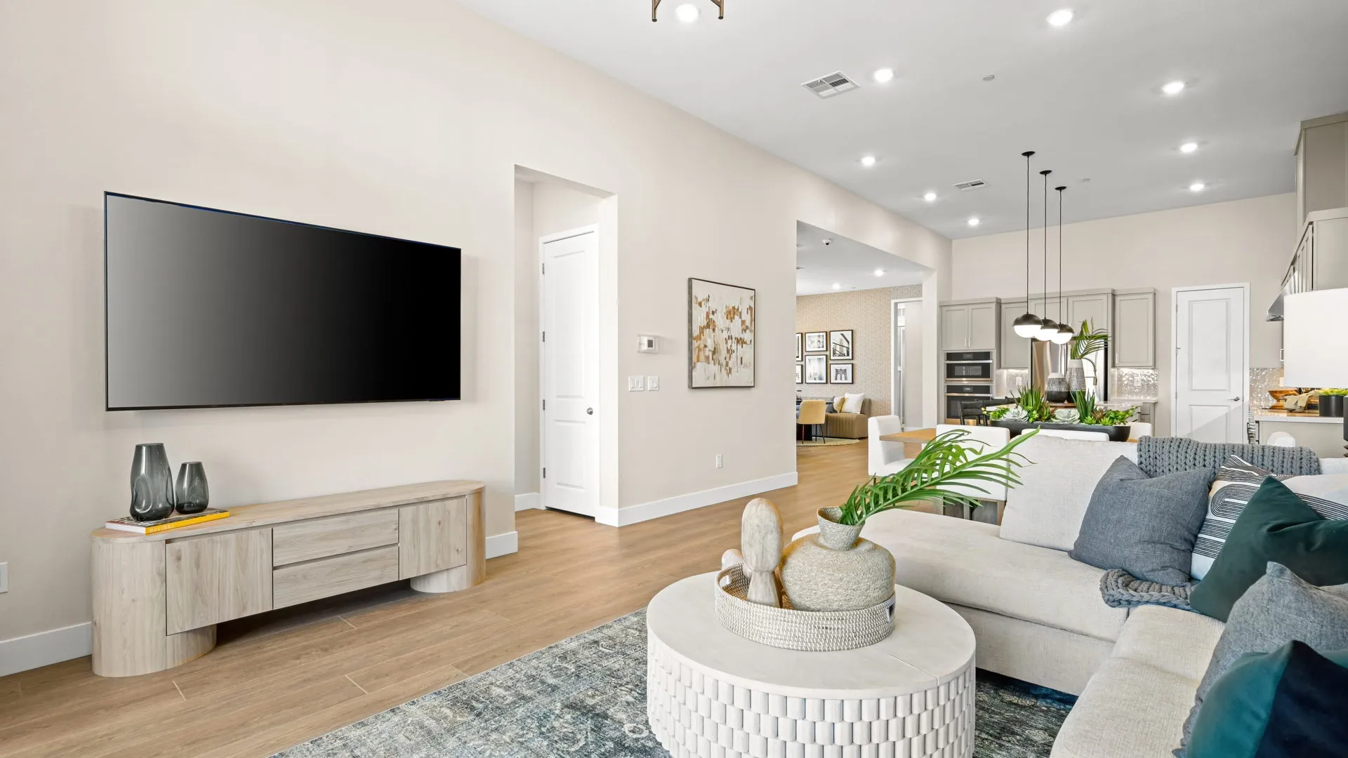 Living Room of Zinnia Model at Esplanade at Red Rock by Taylor Morrison (2) Modern living room with a wall-mounted TV, light wood furniture, a round coffee table with decorative vases, a gray sectional sofa, and an open view into the kitchen and dining area.