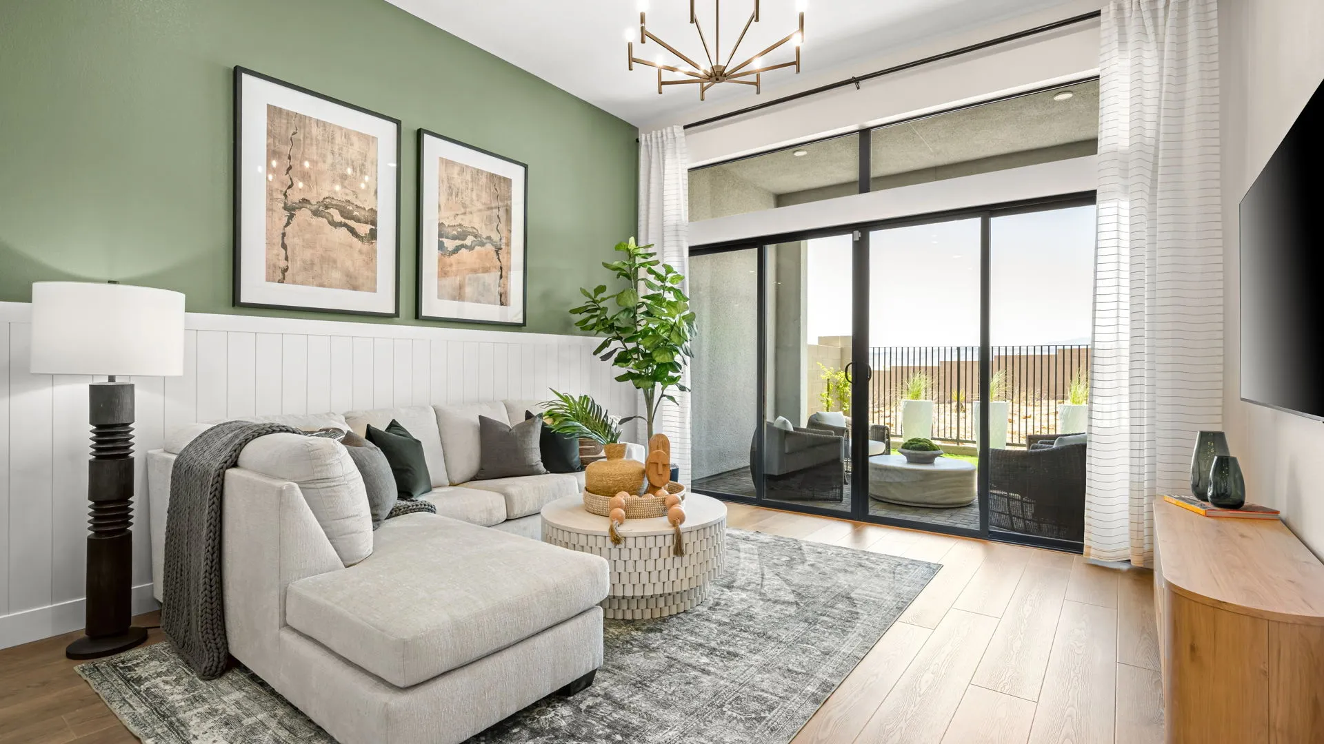 Living Room of Zinnia Model at Esplanade at Red Rock by Taylor Morrison Modern living room with light gray sectional sofa, round ottoman, green accent wall, two abstract art pieces, floor lamp, potted plant, large windows with sheer curtains, wood floors, and a TV on the right wall.