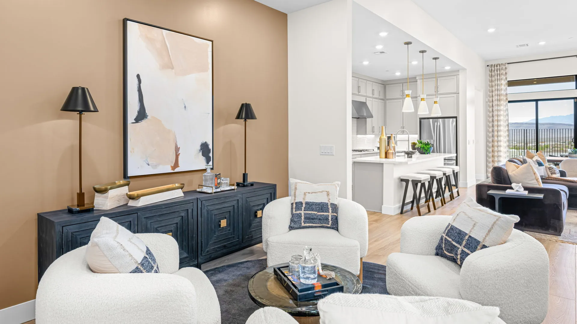 Lounge of Paloma Model at Esplanade at Red Rock by Taylor Morrison (1) Modern living room with four white chairs, a black console with lamps, abstract wall art, and view into a kitchen with white cabinets, an island with barstools, and large windows letting in natural light.