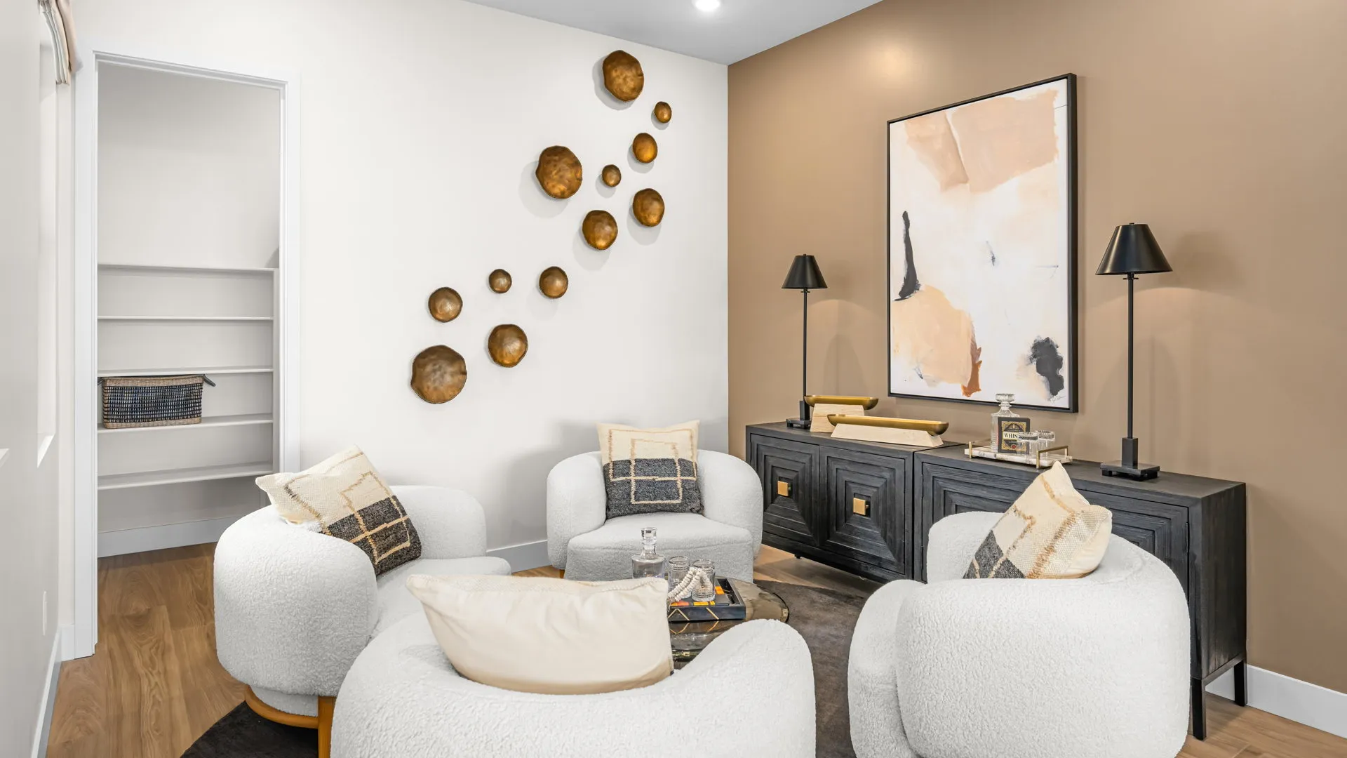 Lounge of Paloma Model at Esplanade at Red Rock by Taylor Morrison Modern living room with four white rounded chairs, a black sideboard with lamps, abstract wall art, and decorative gold wall accents. Shelves are visible in an open nook to the left. Neutral, cozy, and stylish decor.
