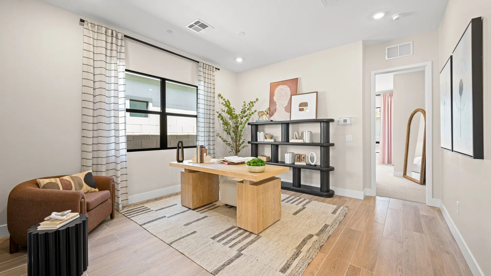 Office of Sangria Model at Esplanade at Red Rock by Taylor Morrison Modern home office with a wooden desk, brown chair, patterned rug, black shelving, framed art, potted plant, and large window with striped curtains, letting in natural light. Doorway leads to another room with a floor mirror.