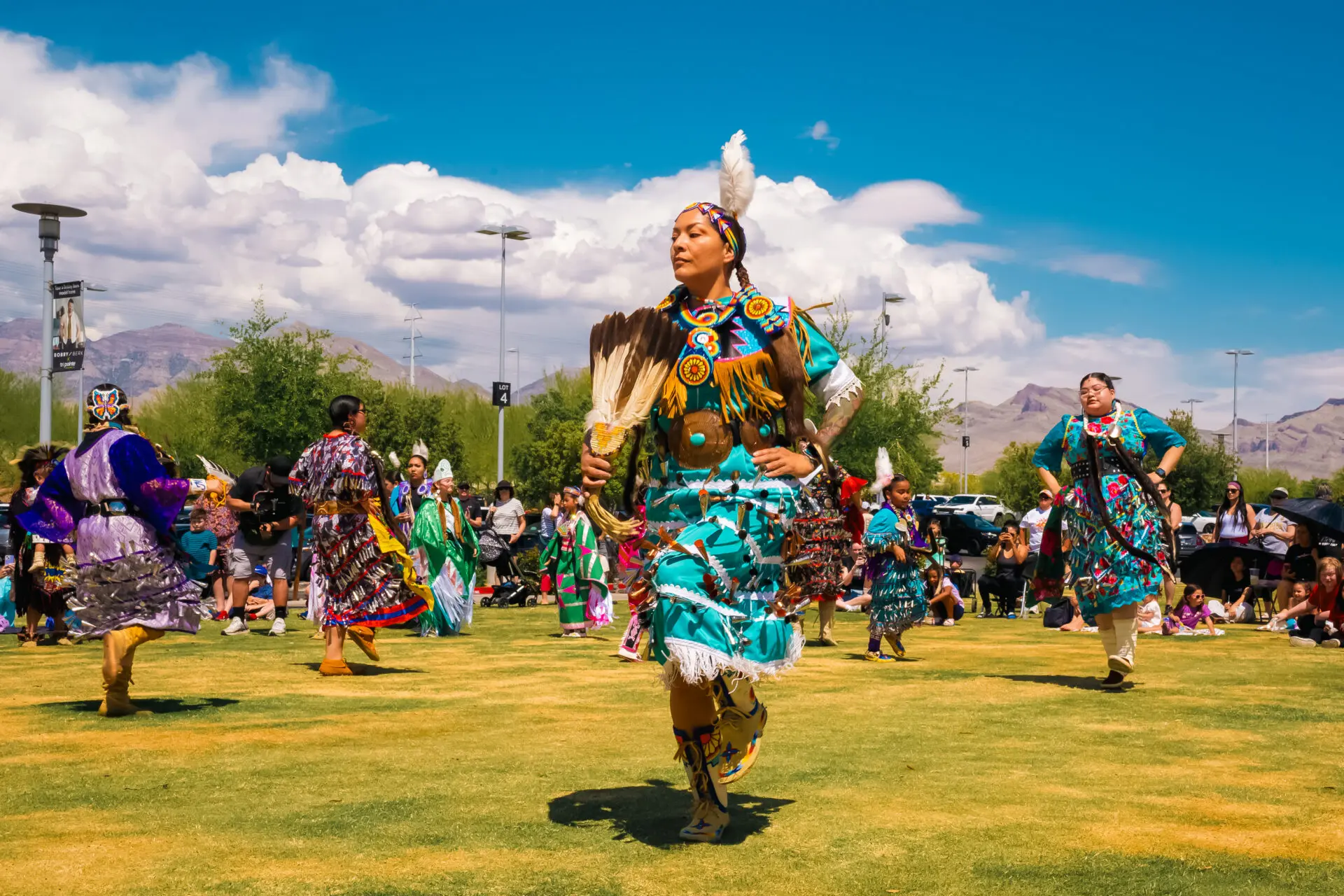 A group of people in colorful traditional Native American regalia dance outdoors on grass under a bright blue sky, with onlookers and mountains visible in the background.