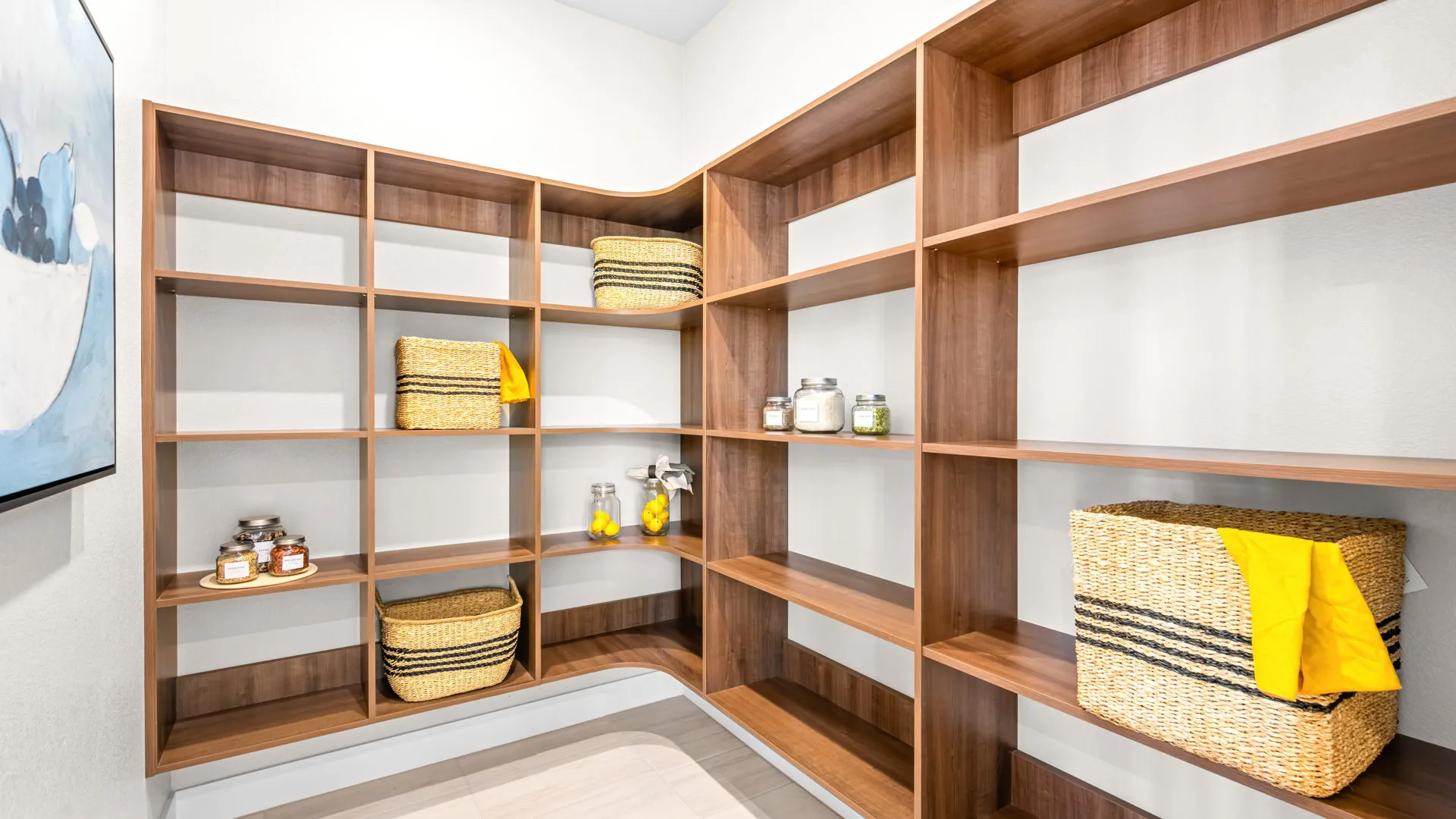 Pantry of Tiki Model at Esplanade at Red Rock by Taylor Morrison Spacious, modern pantry with empty wooden shelves, a few wicker baskets, glass jars, and a folded yellow cloth, set against light-colored walls and floor.