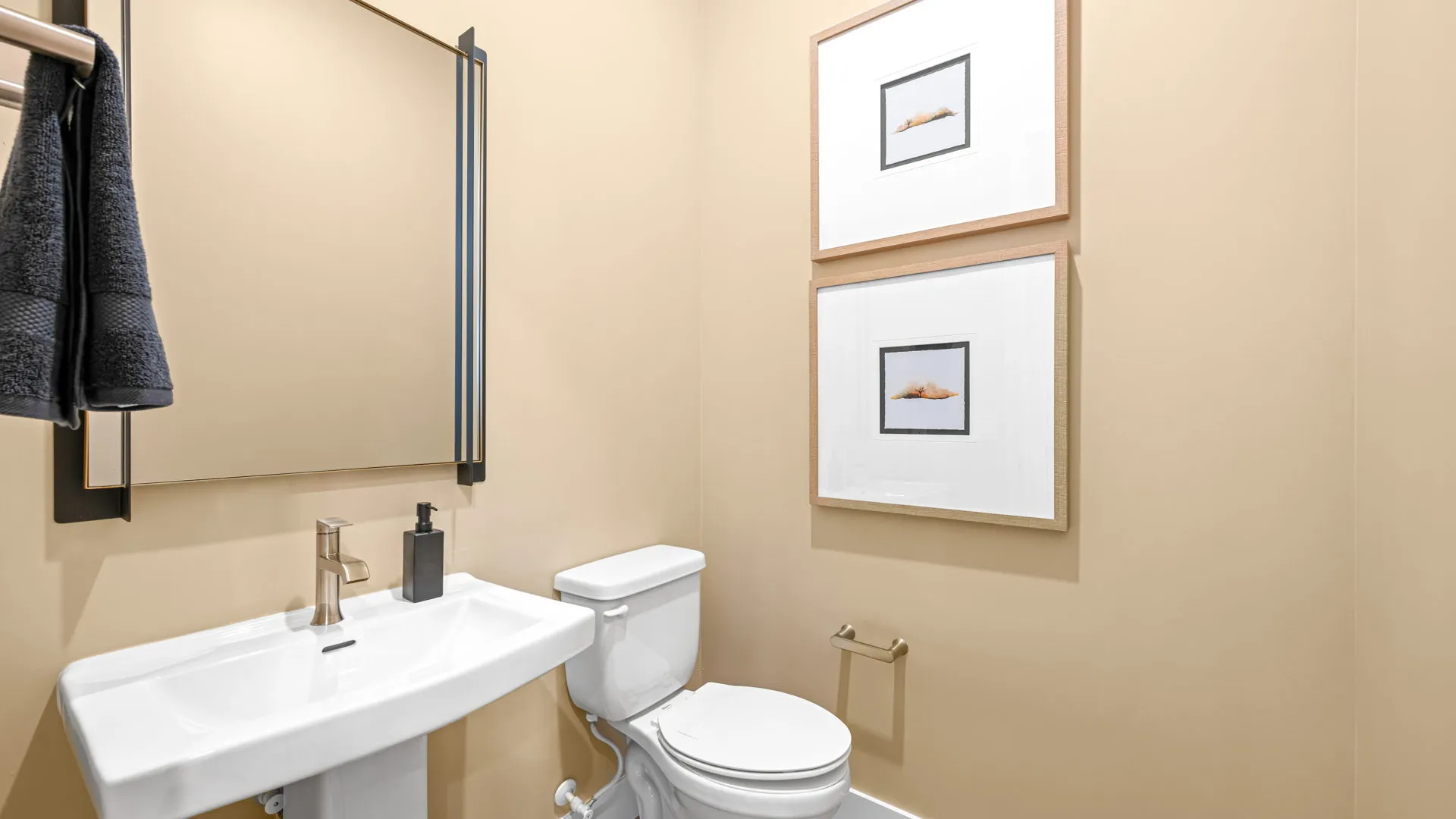 Powder Room of Paloma Model at Esplanade at Red Rock by Taylor Morrison A modern bathroom with beige walls, a white pedestal sink, a white toilet, a rectangular mirror, two framed pictures, and a towel hanging from a rack.
