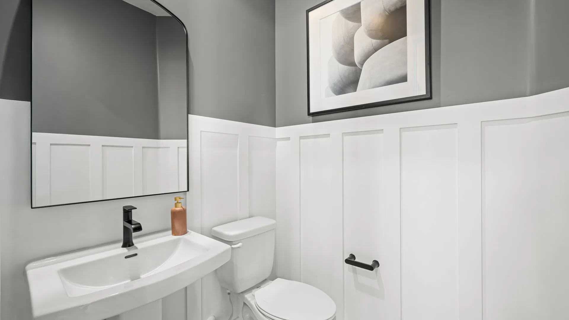 Powder Room of Sangria Model at Esplanade at Red Rock by Taylor Morrison A modern bathroom with white wainscoting, gray upper walls, a rectangular mirror, a white sink with black faucet, a toilet, soap dispenser, toilet paper holder, and abstract artwork above the toilet.