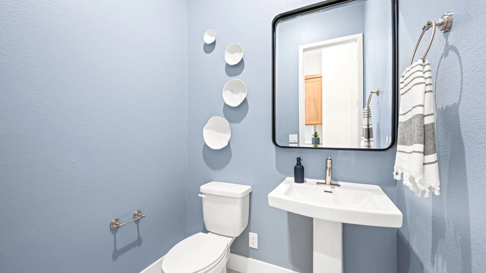 Powder Room of Tiki Model at Esplanade at Red Rock by Taylor Morrison A modern bathroom with light blue walls, a pedestal sink, a toilet, a towel ring with a striped towel, wall-mounted decor plates, and a large mirror reflecting a door and cabinet.