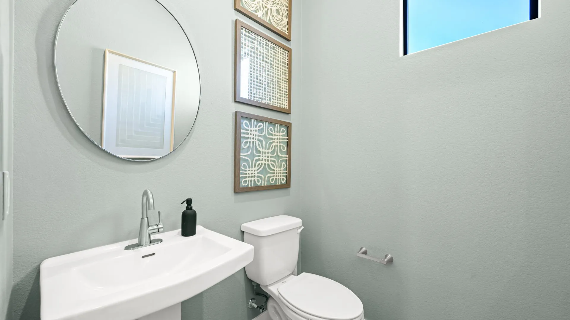Powder Room of Zinnia Model at Esplanade at Red Rock by Taylor Morrison A modern bathroom with a round mirror, pedestal sink, black soap dispenser, white toilet, wall art, and a small rectangular window.