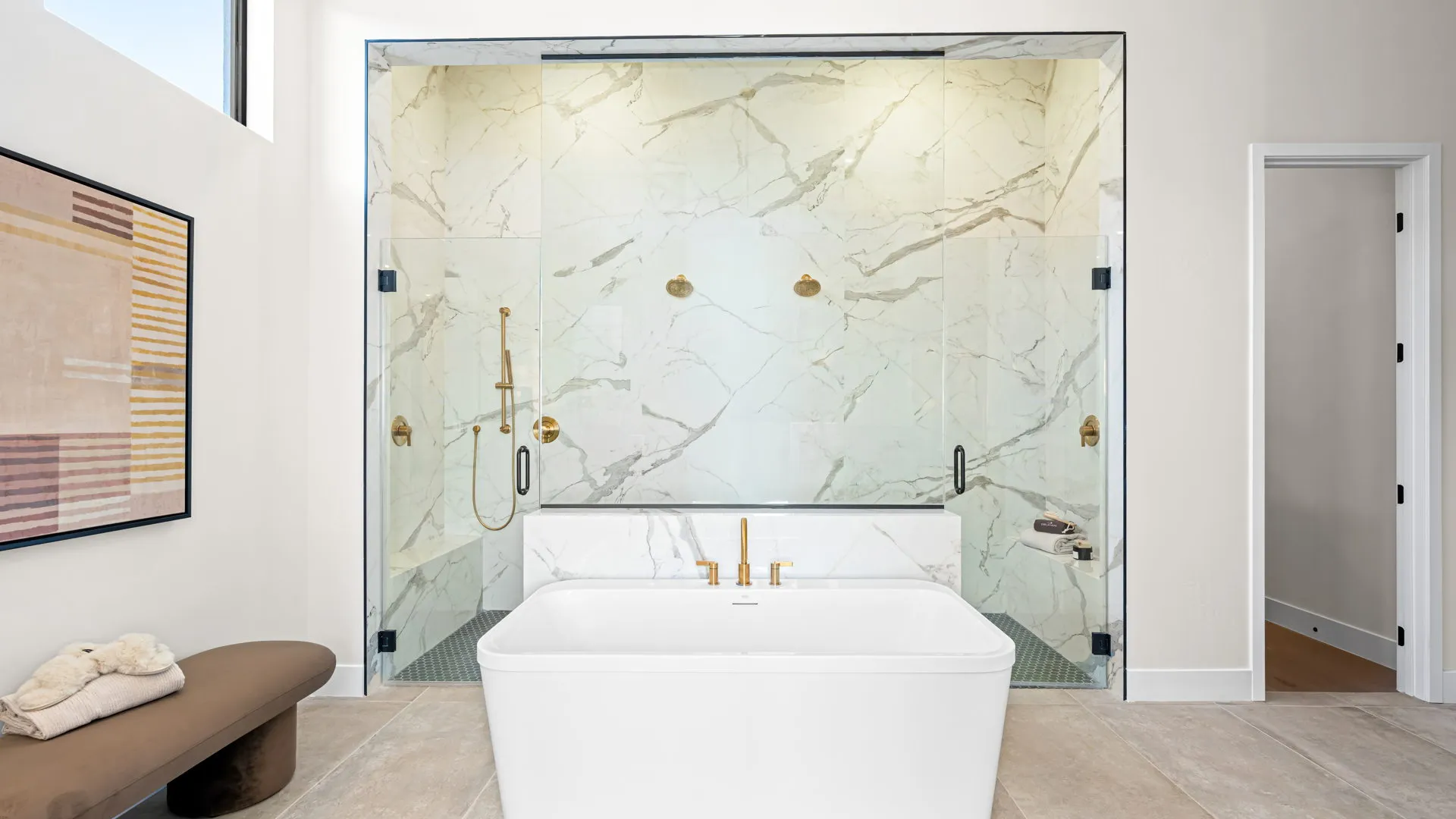 Primary Bathrom of Monroe Model at Esplanade at Red Rock by Taylor Morrison Modern bathroom with a freestanding white tub, gold fixtures, and a large shower with marble walls and glass doors. A brown bench with towels and a framed abstract artwork are also visible.