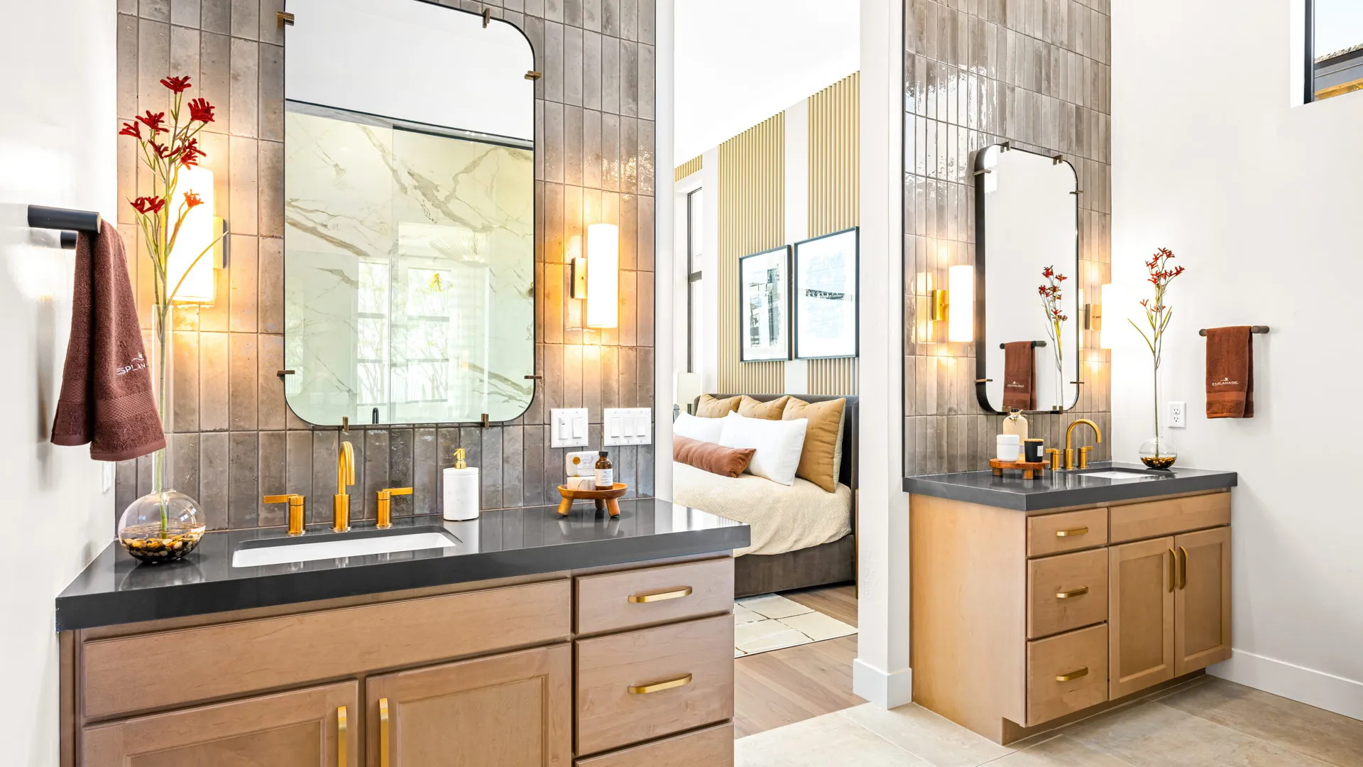 Primary Bathroom of Monroe Model at Esplanade at Red Rock by Taylor Morrison (1) Modern bathroom with two wooden vanities, gold fixtures, large mirrors, wall sconces, and tiled walls. Reflected in the mirror is a glimpse of a cozy bedroom with framed art on the wall.