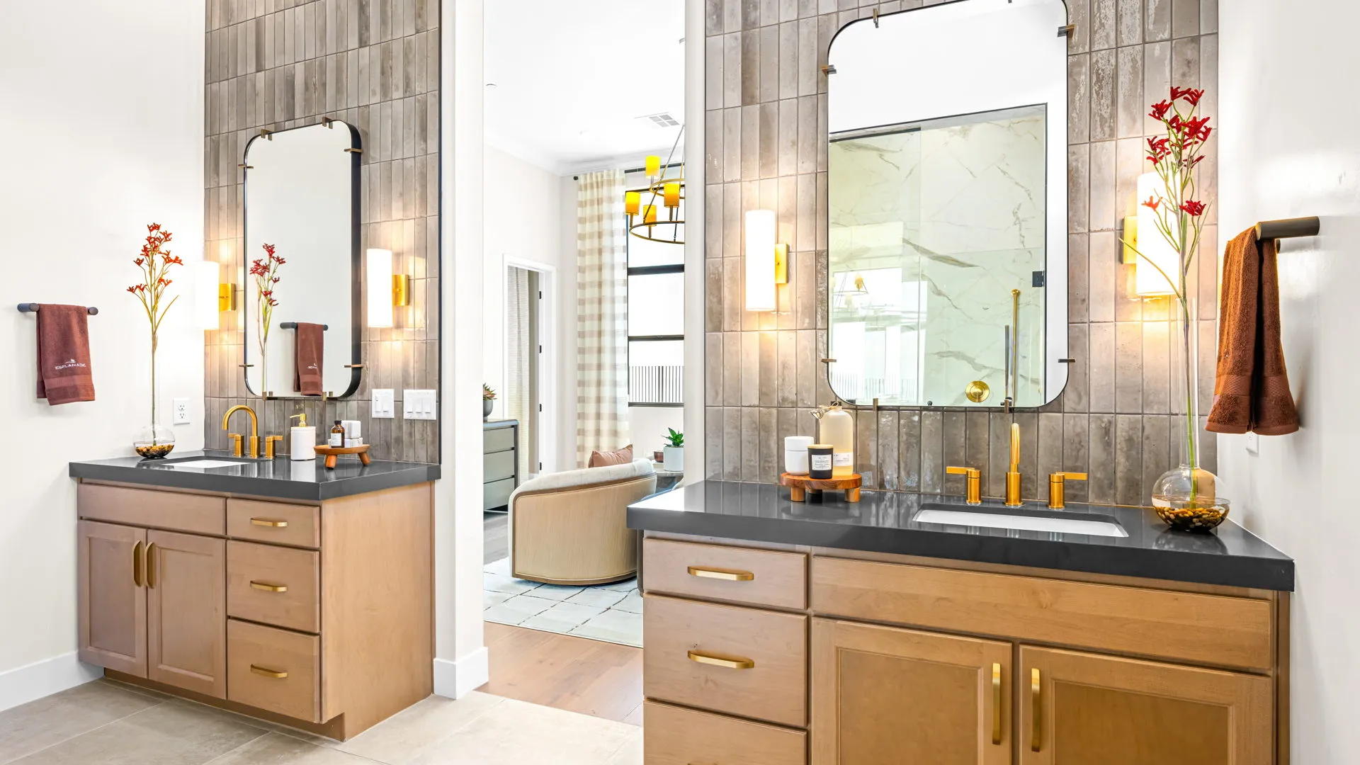 Primary Bathroom of Monroe Model at Esplanade at Red Rock by Taylor Morrison (2) Modern bathroom with two wood vanities, black countertops, large mirrors, gold fixtures, wall sconces, brown towels, and red flower accents. A bright room with a chair is visible through the doorway.