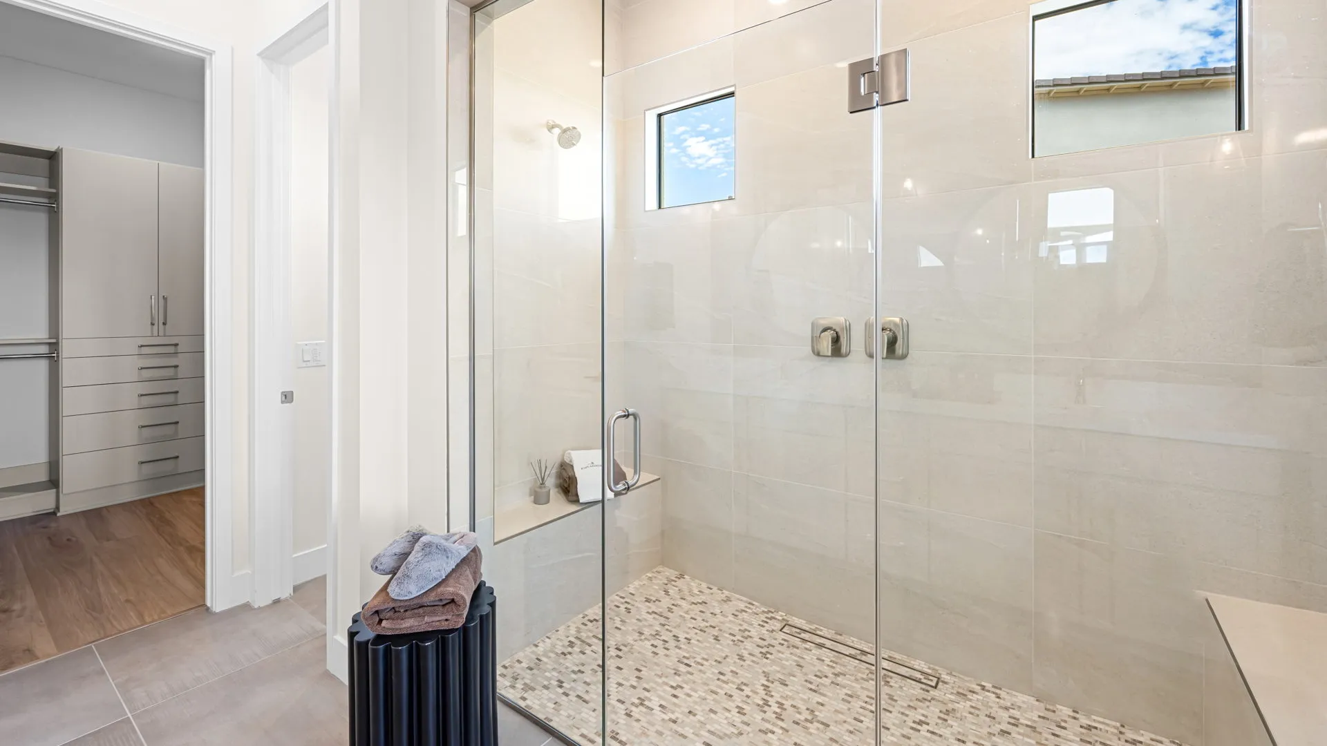 Primary Bathroom of Paloma Model at Esplanade at Red Rock by Taylor Morrison (1) Modern bathroom with a large glass-enclosed shower, beige tiles, built-in bench, and two small windows. Outside the shower, folded towels rest on a black laundry hamper near the entrance to a closet.