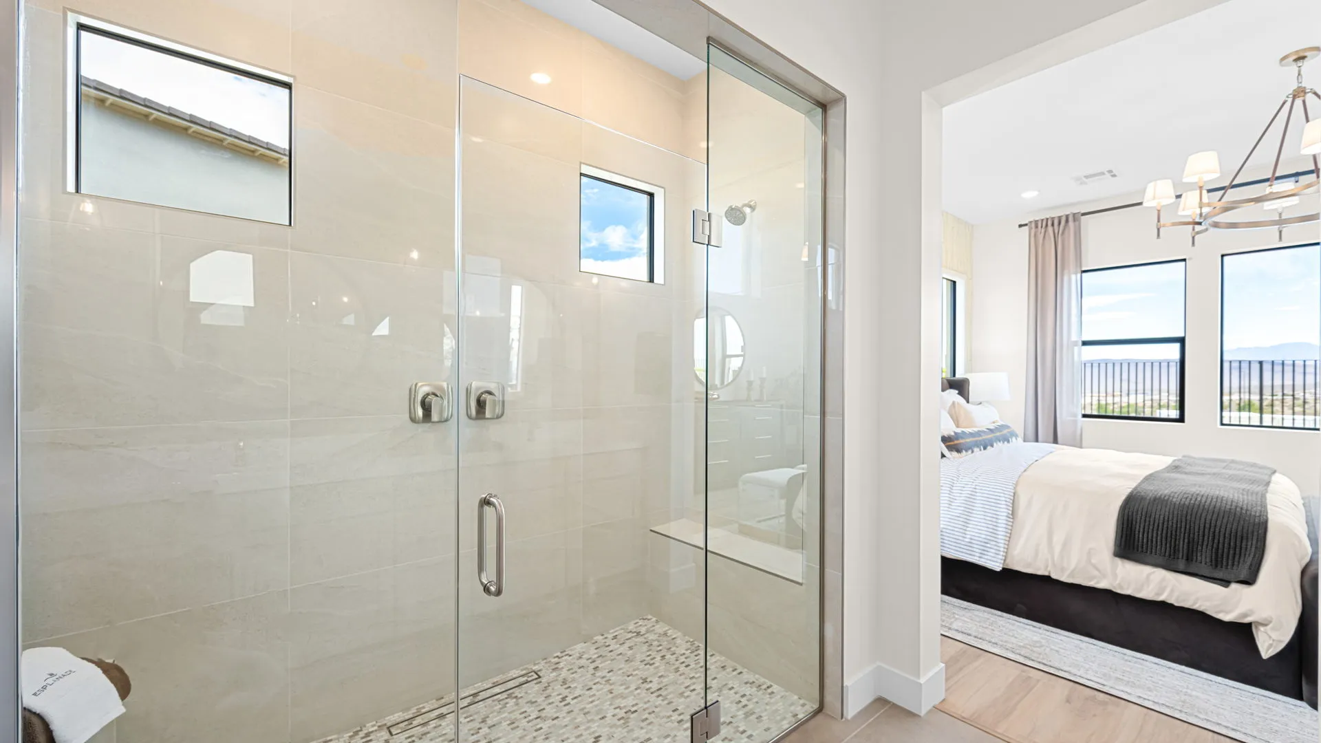 Primary Bathroom of Paloma Model at Esplanade at Red Rock by Taylor Morrison (2) Modern bathroom with a large glass shower featuring dual showerheads and beige tile; adjacent bedroom with a made bed, chandelier, and large windows with views outside.