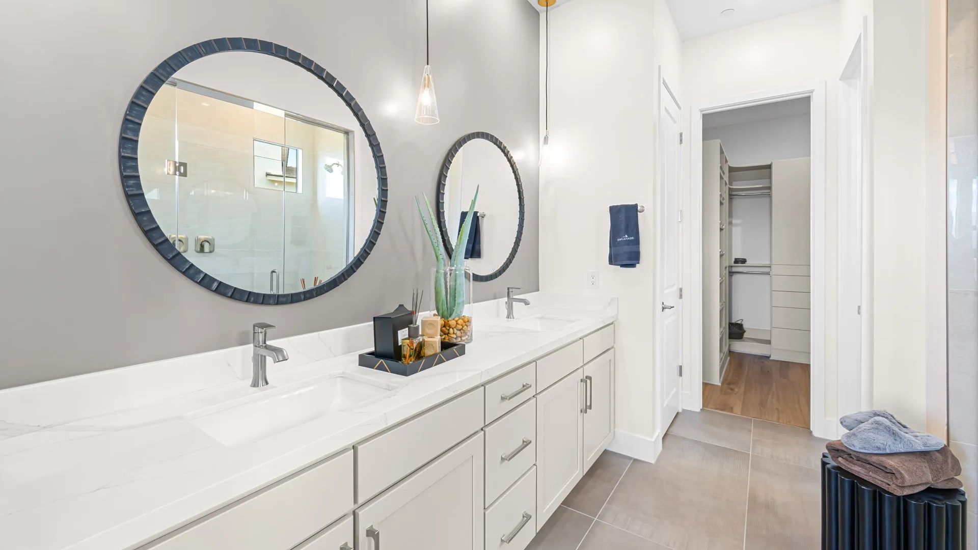 Primary Bathroom of Paloma Model at Esplanade at Red Rock by Taylor Morrison Modern bathroom with double sinks, large round mirrors, white cabinets, a tray with decor items, and a tall plant. There is a walk-in closet visible through an open door in the background.