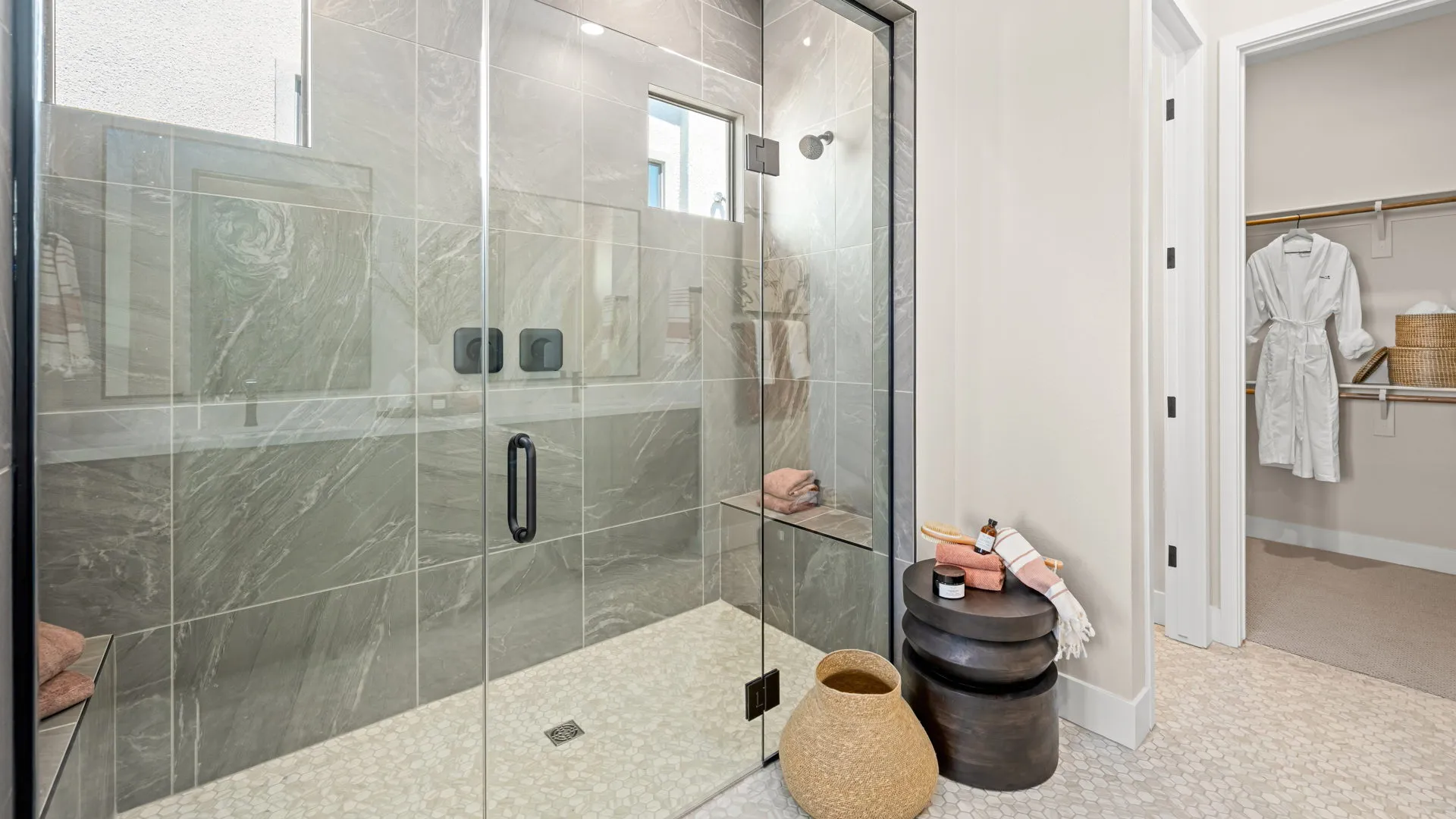 Primary Bathroom of Sangria Model at Esplanade at Red Rock by Taylor Morrison (1) Modern bathroom with a glass shower enclosure, gray tiled walls, two black nested tables with towels and bath items, a woven basket, and a closet with a white robe visible through an open door.