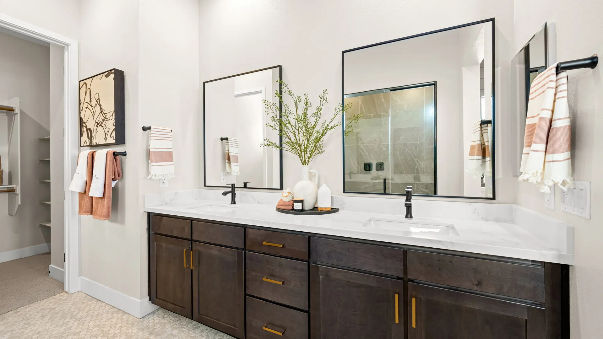 Primary Bathroom of Sangria Model at Esplanade at Red Rock by Taylor Morrison Modern bathroom with dark wood double vanity, two square mirrors, black faucets, and white countertops. Orange and white towels hang on black bars, and a vase with branches decorates the counter. A glass shower is visible in the background.