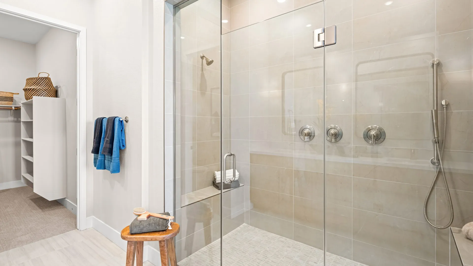 Primary Bathroom of Tiki Model at Esplanade at Red Rock by Taylor Morrison (1) Modern bathroom with a large glass-enclosed shower featuring three handles, beige tile walls, a bench, and a wooden stool with a brush and soap. Towels hang on the wall and an adjacent walk-in closet is visible.
