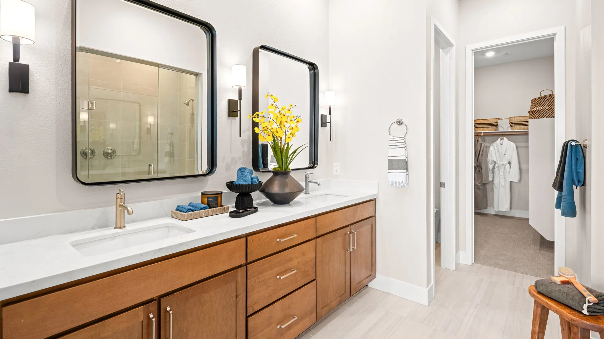 Primary Bathroom of Tiki Model at Esplanade at Red Rock by Taylor Morrison Modern bathroom with two sinks, large mirrors, wooden cabinets, and a vase of yellow flowers. Towels and toiletries are neatly arranged, and a walk-in closet with robes and shelves is visible in the background.