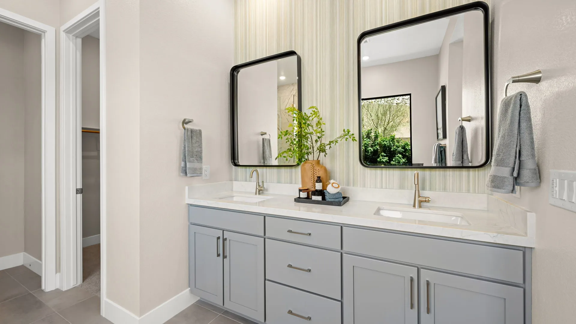 Primary Bathroom of Zinnia Model at Esplanade at Red Rock by Taylor Morrison (1) Modern bathroom with dual sinks, gray cabinets, two mirrors, gold faucets, towel racks, decorative tray with toiletries, and a plant. A window shows greenery outside. Neutral wall colors and tile floor.