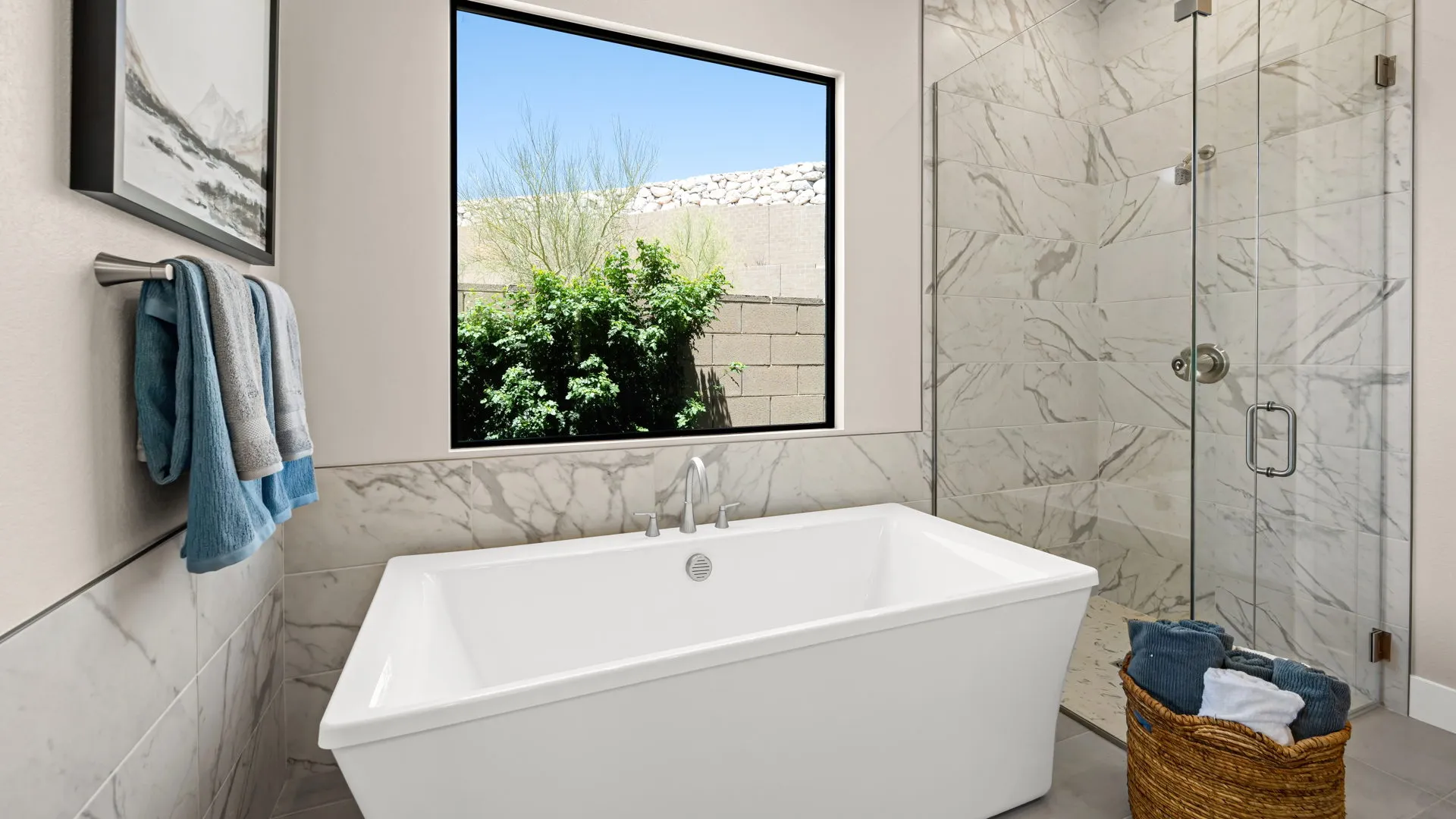 Primary Bathroom of Zinnia Model at Esplanade at Red Rock by Taylor Morrison Modern bathroom with a rectangular white bathtub, marble-patterned walls, glass shower, towels on a rack, a basket with towels, and a large window overlooking a green shrub and brick wall outside.