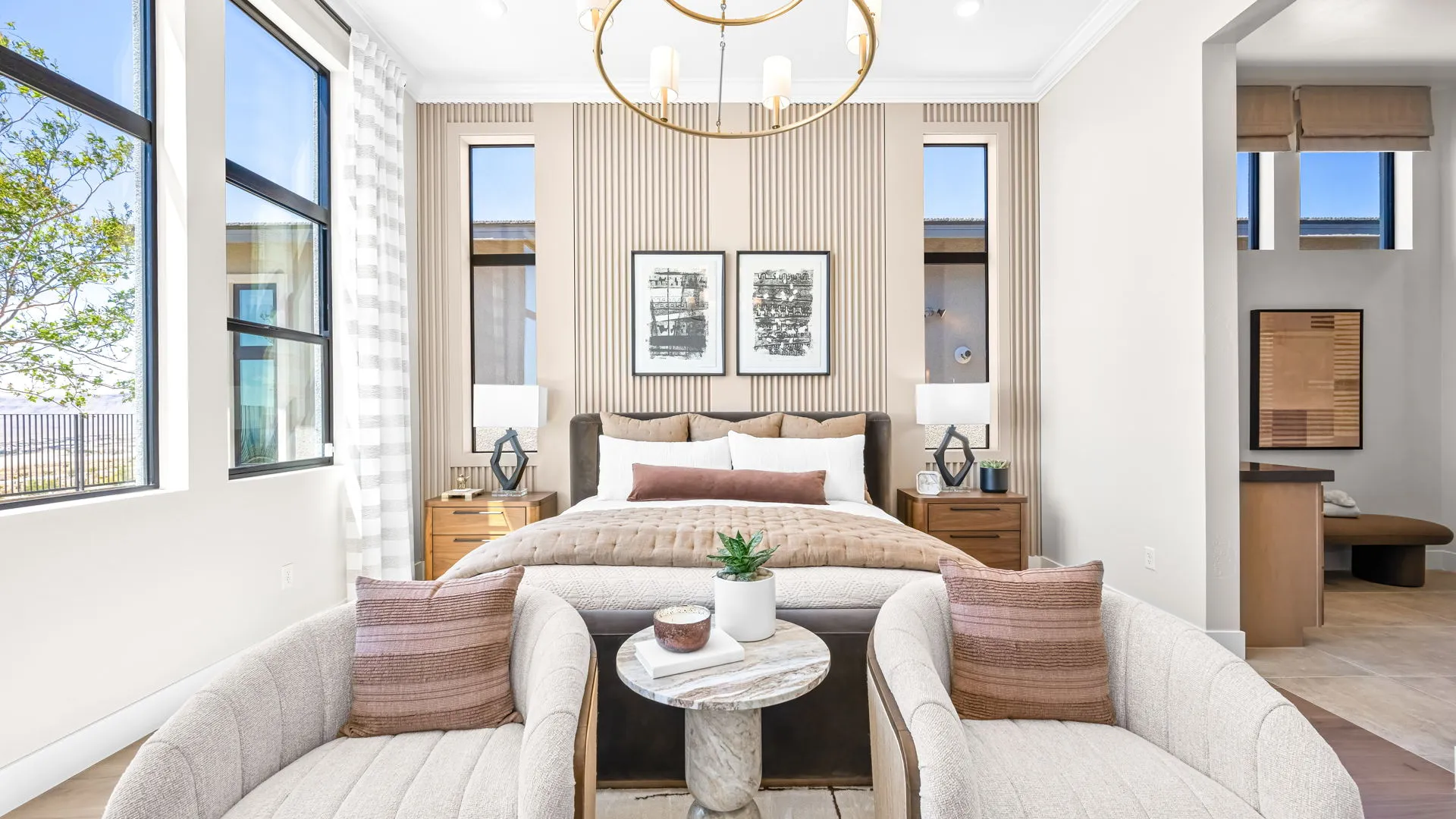 Primary Bedroom of Monroe Model at Esplanade at Red Rock by Taylor Morrison (2) Bright modern bedroom with a bed, two framed pictures above, two nightstands with lamps, two beige chairs, a round marble table, and large windows letting in natural light.