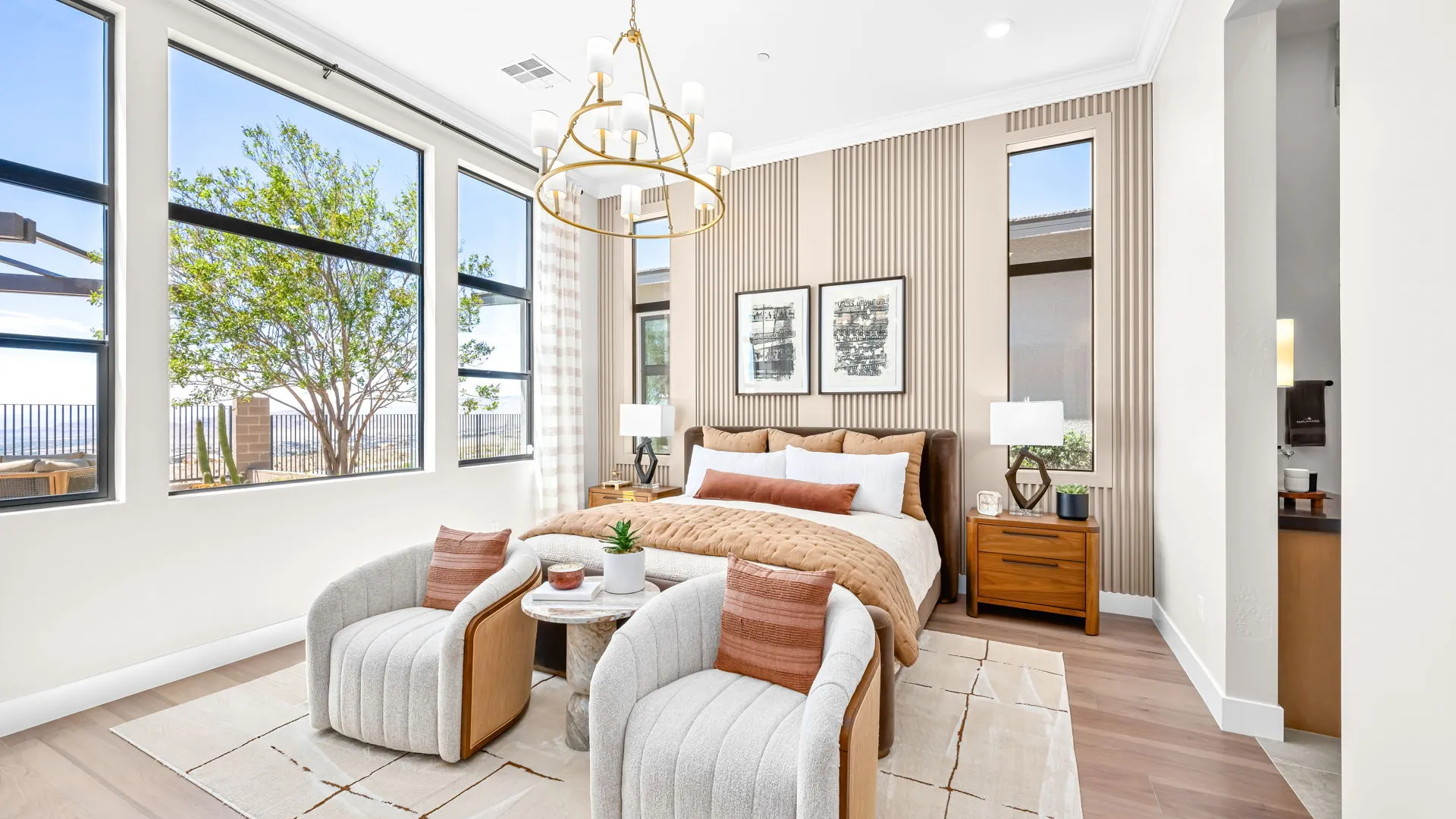 Primary Bedroom of Monroe Model at Esplanade at Red Rock by Taylor Morrison Bright modern bedroom with a large bed, two cushioned chairs, a chandelier, and big windows letting in natural light. Neutral tones and wood accents create a cozy, elegant atmosphere.