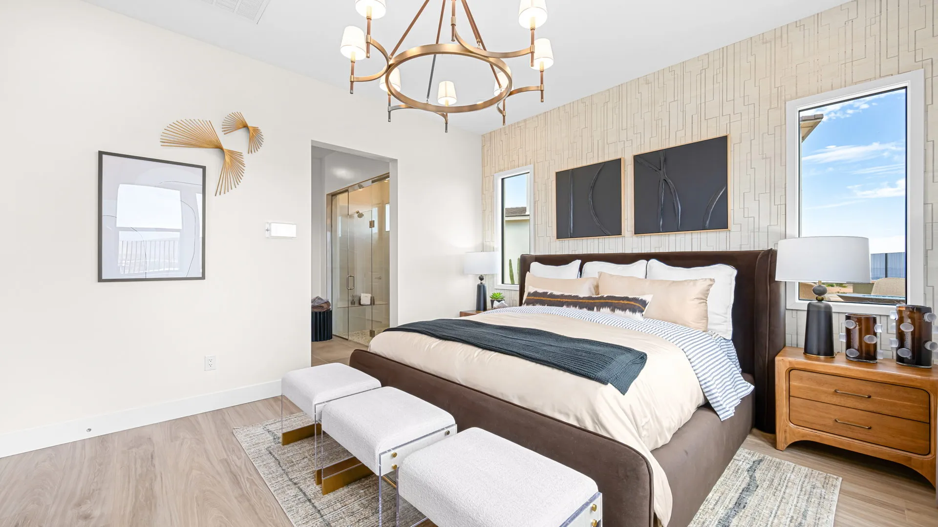 Primary Bedroom of Paloma Model at Esplanade at Red Rock by Taylor Morrison (1) Modern bedroom with a large bed, two windows, abstract wall art, a chandelier, two small benches, a nightstand with decor, and an open door leading to a bathroom.