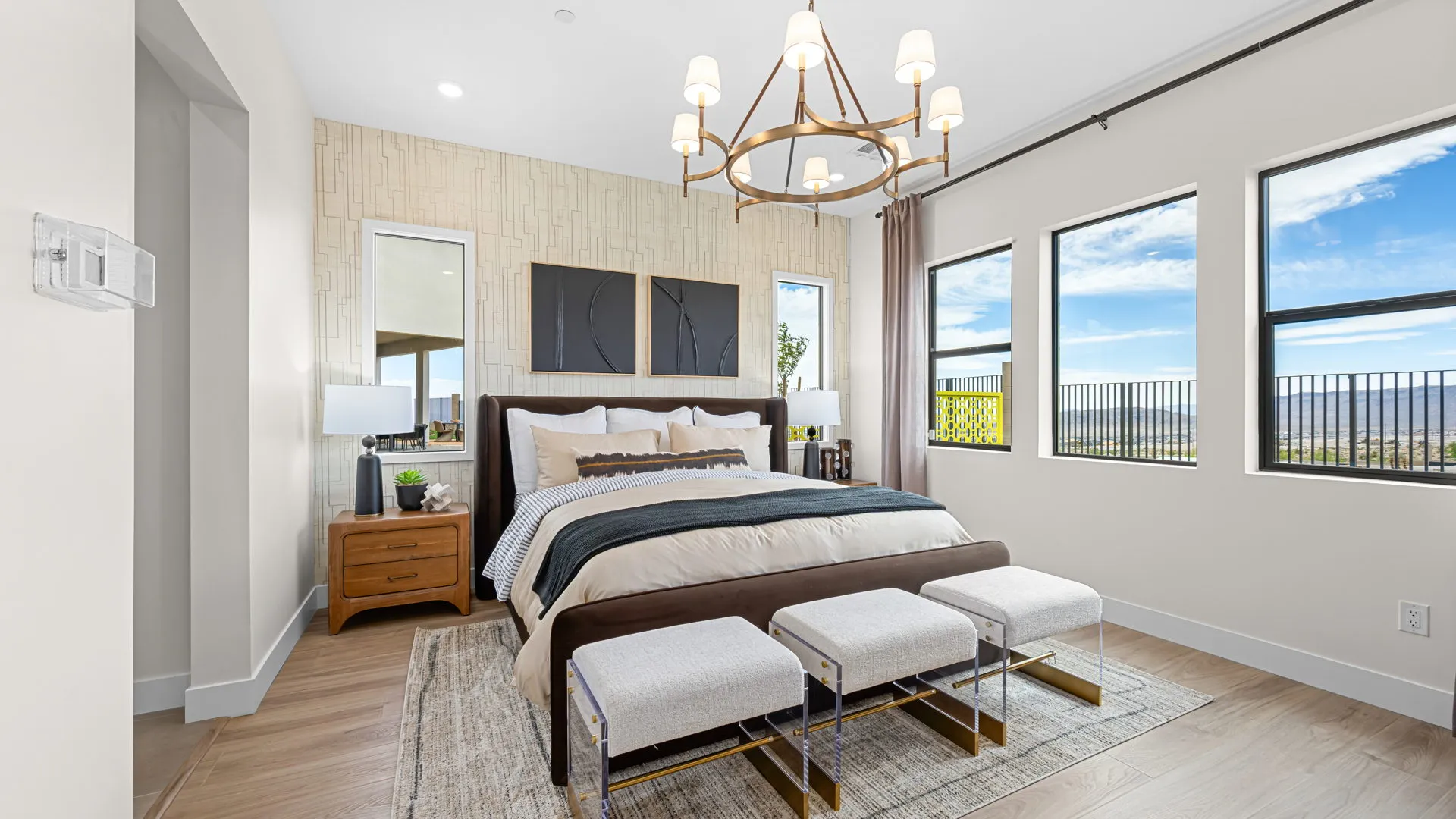 Primary Bedroom of Paloma Model at Esplanade at Red Rock by Taylor Morrison Modern bedroom with a large bed, two nightstands, three benches at the foot, and a chandelier. Light walls, wood floor, and three windows showing an outdoor view with blue sky and clouds.