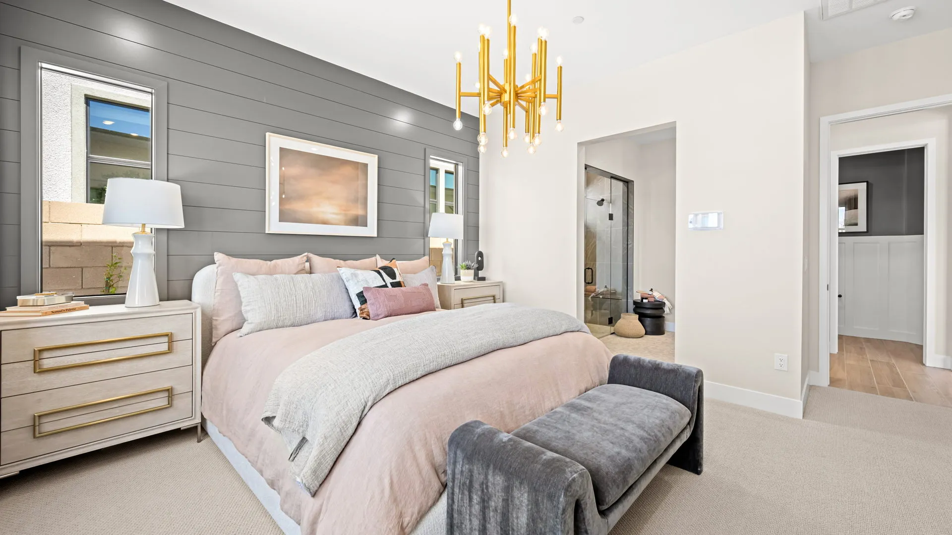 Primary Bedroom of Sangria Model at Esplanade at Red Rock by Taylor Morrison (1) Modern bedroom with a beige bed, gray accent wall, two windows, gold chandelier, bedside tables with lamps, and a bench at the foot of the bed. A doorway reveals a bathroom with a glass shower.