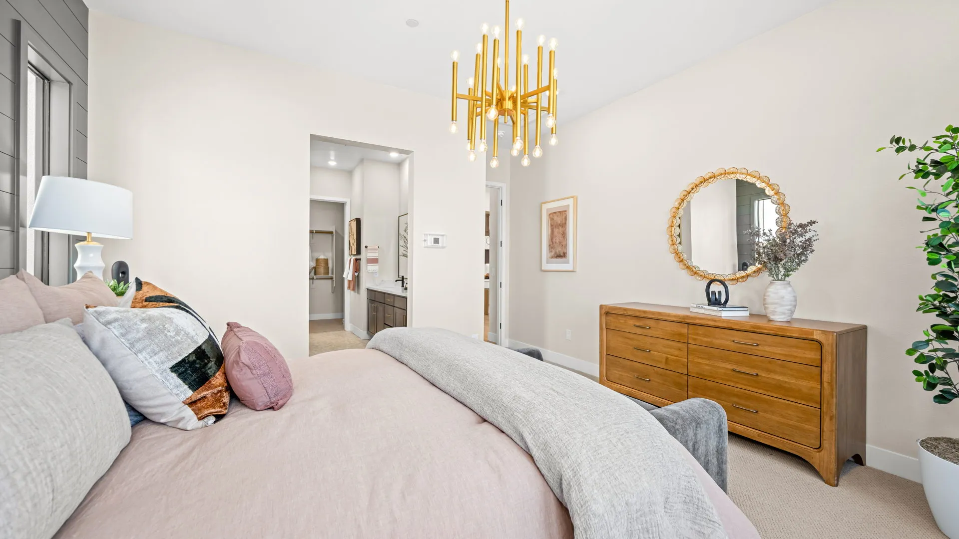 Primary Bedroom of Sangria Model at Esplanade at Red Rock by Taylor Morrison (2) Bright bedroom with a modern gold chandelier, beige walls, a bed with neutral bedding, a wooden dresser with a round mirror, a gray bench, and a potted plant in the corner. Doorway leads to another room.