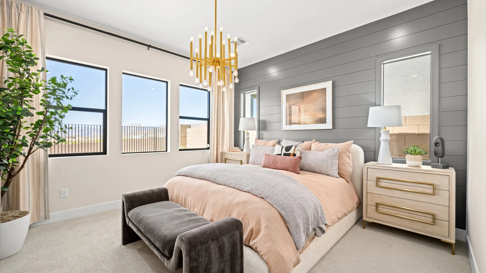 Primary Bedroom of Sangria Model at Esplanade at Red Rock by Taylor Morrison Modern bedroom with a large bed, pink and gray bedding, gold chandelier, two nightstands with lamps, wall art, indoor plant, and windows letting in natural light.