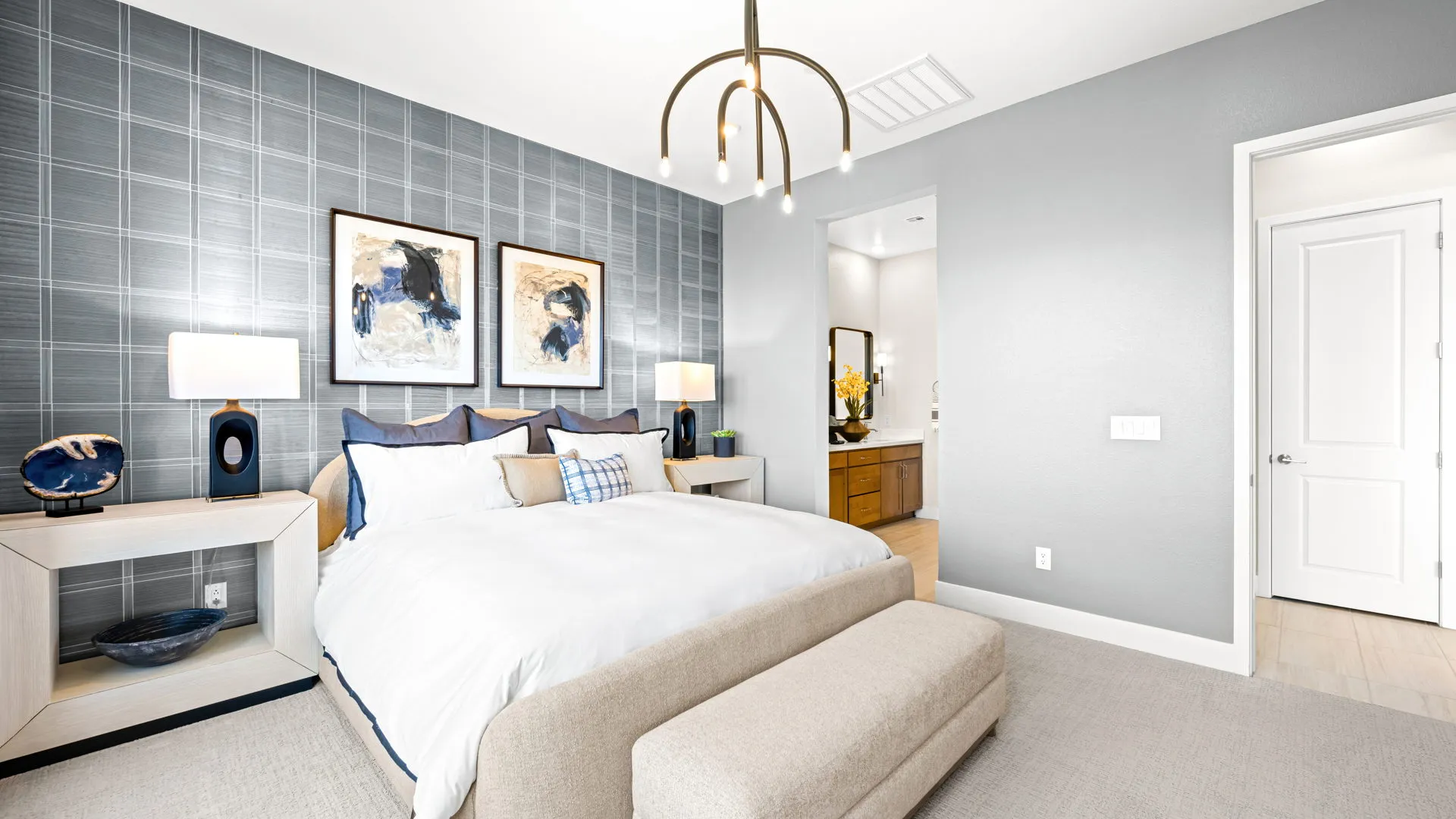 Primary Bedroom of Tiki Model at Esplanade at Red Rock by Taylor Morrison (1) Modern bedroom with a large bed, white bedding, abstract artwork above the headboard, two nightstands with lamps, a light fixture overhead, and an open doorway leading to a bathroom.