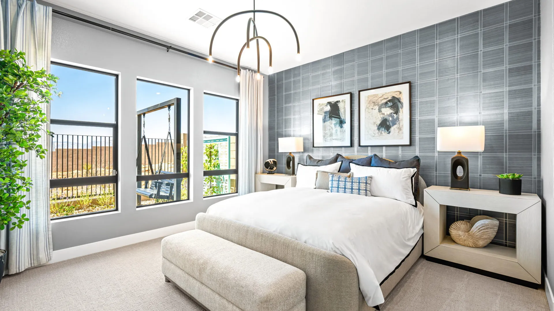 Primary Bedroom of Tiki Model at Esplanade at Red Rock by Taylor Morrison Modern bedroom with large windows, gray accent wall, abstract art, two lamps on nightstands, a white bed, gray bench, and a potted plant by the window. Natural light fills the room.