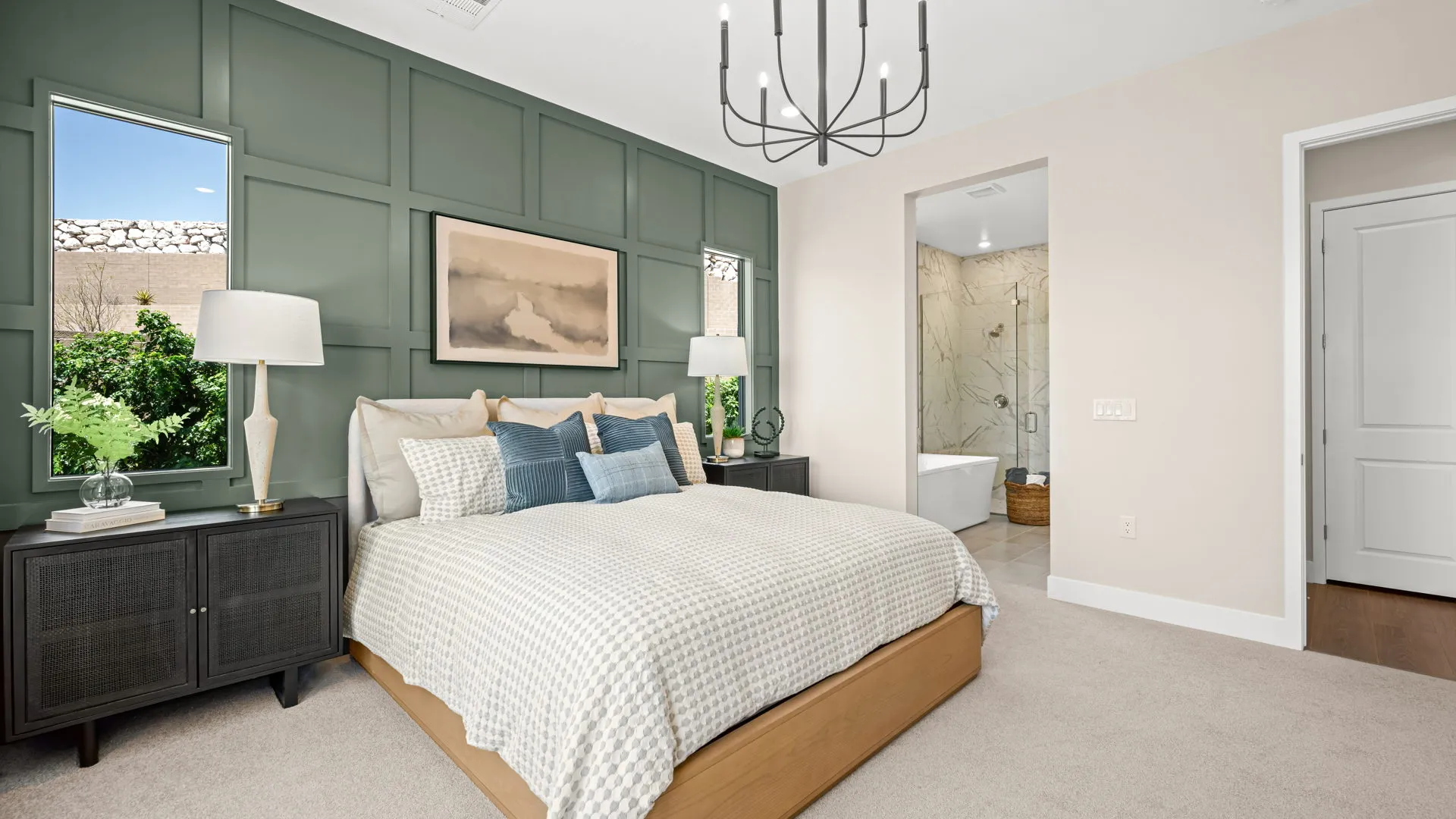 Primary Bedroom of Zinnia Model at Esplanade at Red Rock by Taylor Morrison (1) Modern bedroom with a green accent wall, beige bed, blue and white pillows, two dark nightstands with lamps, large artwork, and an ensuite bathroom with a bathtub visible through an open doorway.