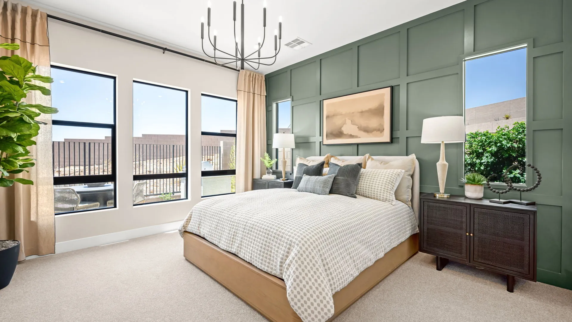 Primary Bedroom of Zinnia Model at Esplanade at Red Rock by Taylor Morrison A modern bedroom with green accent wall, large bed with patterned bedding, two side tables with lamps, three tall windows, beige curtains, and a chandelier. A potted plant sits in the corner near the windows.