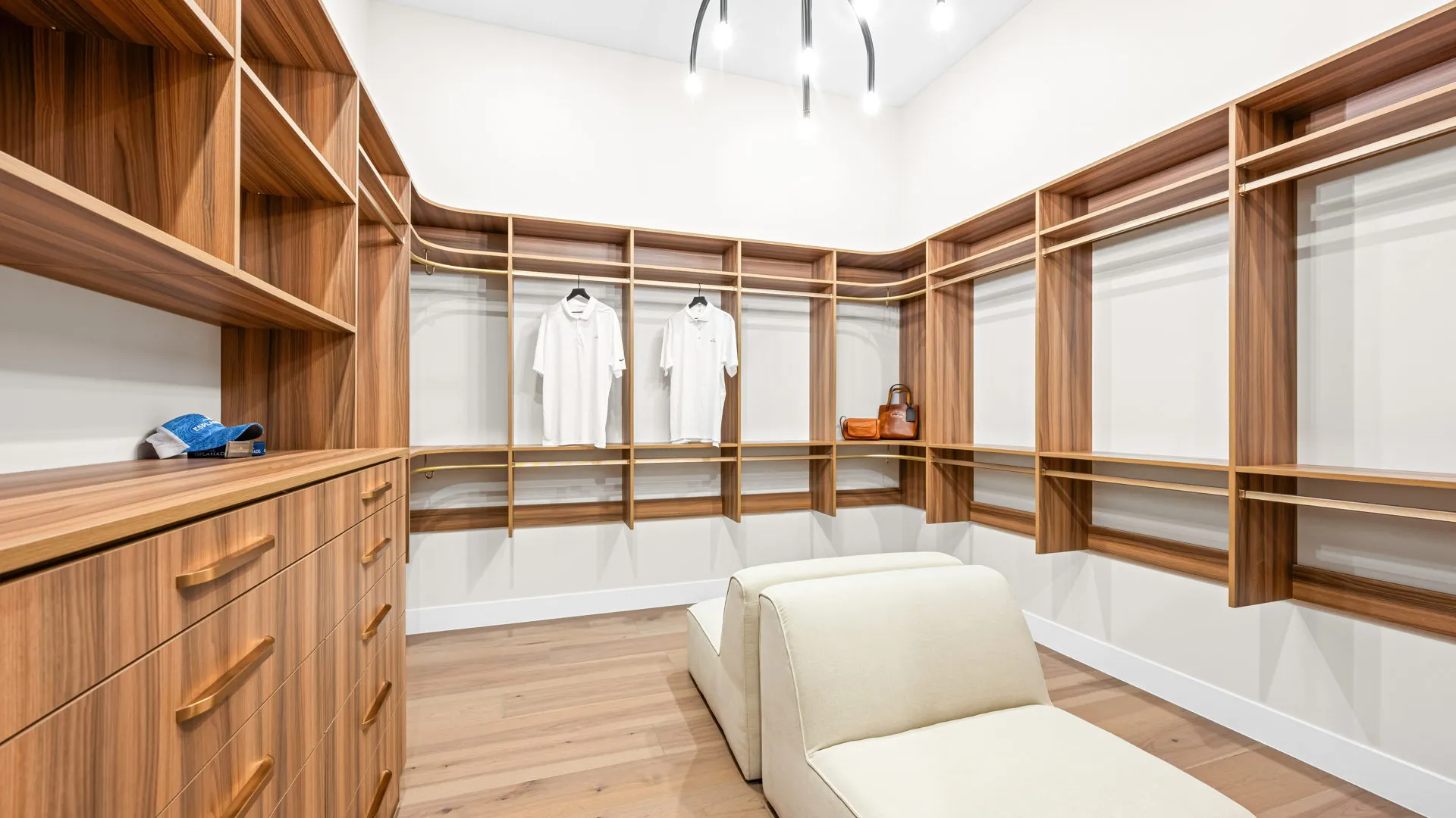 Primary Closet of Monroe Model at Esplanade at Red Rock by Taylor Morrison Spacious walk-in closet with wooden shelves, drawers, two white shirts on hangers, a pair of shoes, two handbags, and a cream-colored bench under modern ceiling lights.