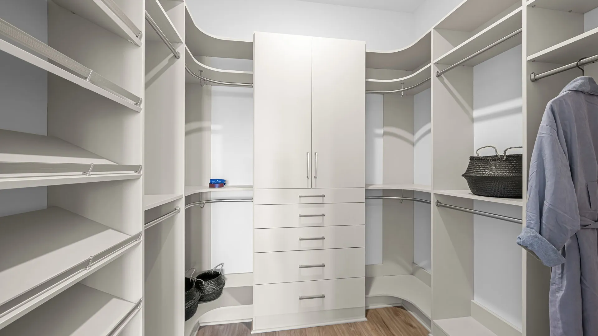 Primary Closet of Paloma Model at Esplanade at Red Rock by Taylor Morrison Spacious walk-in closet with white shelves, drawers, and hanging space. Two baskets sit on shelves, a robe hangs on the right, and a small box is on the left shelf. Light wood flooring.