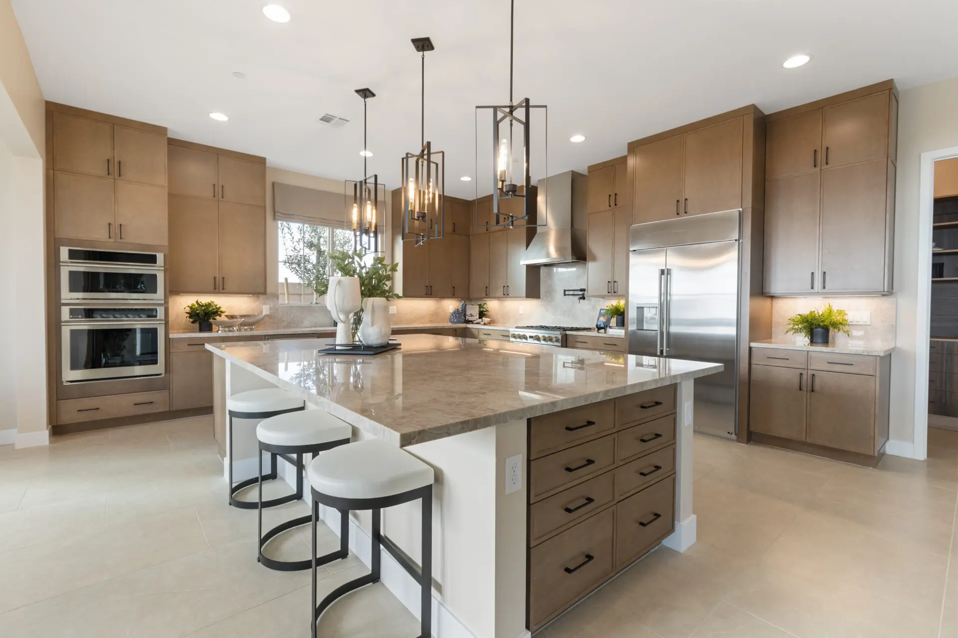 Modern kitchen with light brown cabinets, marble island with three white stools, pendant lights, stainless steel appliances, built-in ovens, and decorative plants on counters. Spacious, bright, and neatly organized.