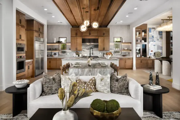 Modern open-concept kitchen and living area with wood cabinetry, marble island, pendant lights, a set dining table, a white sofa with patterned pillows, decorative accents, and natural light streaming in.