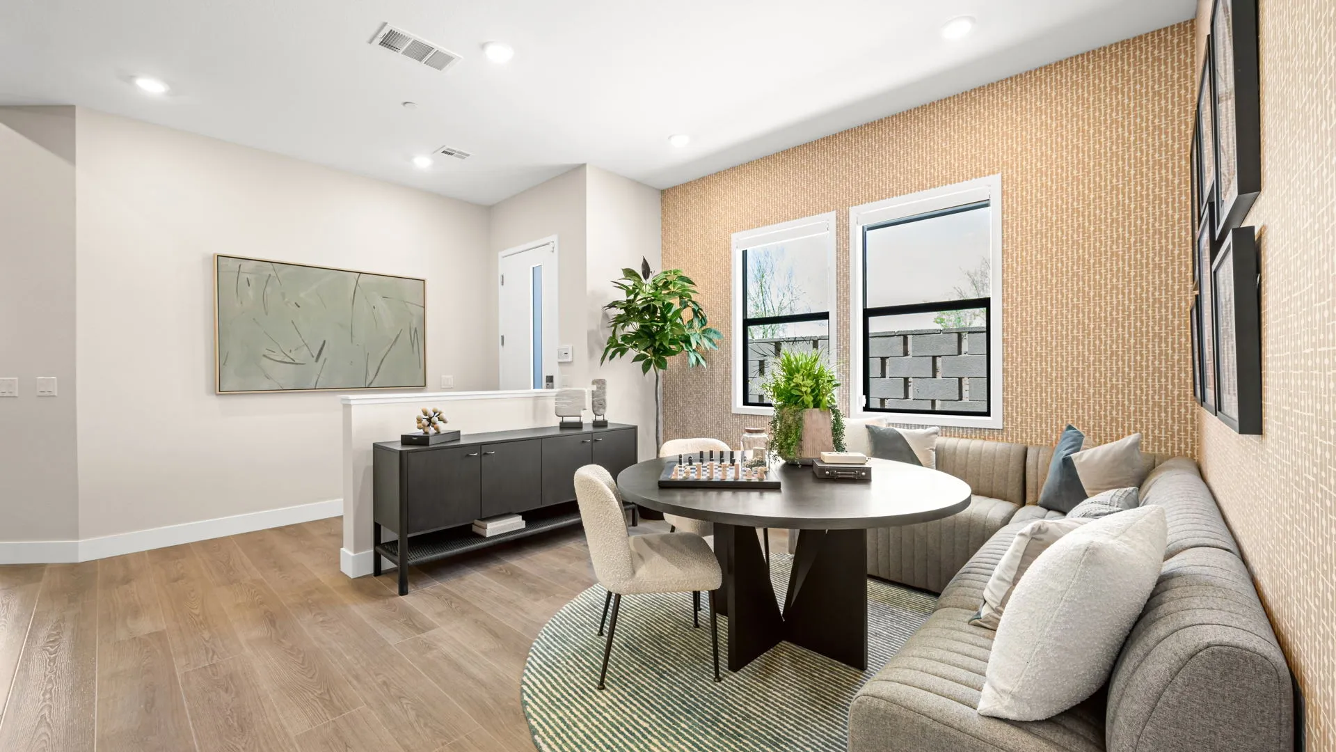 Seating Area of Zinnia Model at Esplanade at Red Rock by Taylor Morrison (1) Modern living room with wood flooring, a round table with chairs, a light gray sofa, green plant, console table, abstract artwork, and windows letting in natural light. Decor is minimalist and stylish.