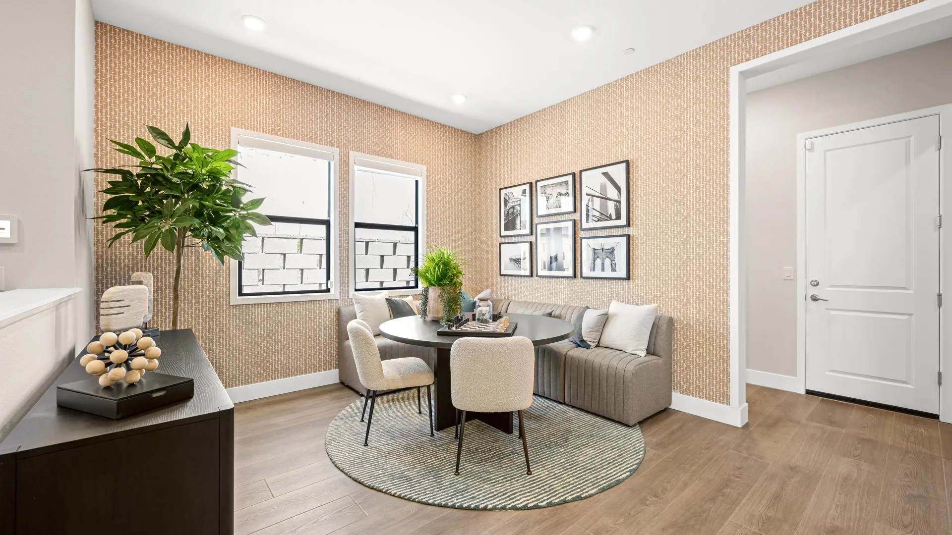 Seating Area of Zinnia Model at Esplanade at Red Rock by Taylor Morrison Modern dining area with a round table, four beige chairs, a gray couch, green plants, wall art, and wood flooring. Two windows let in natural light, and a white door is visible on the right side.
