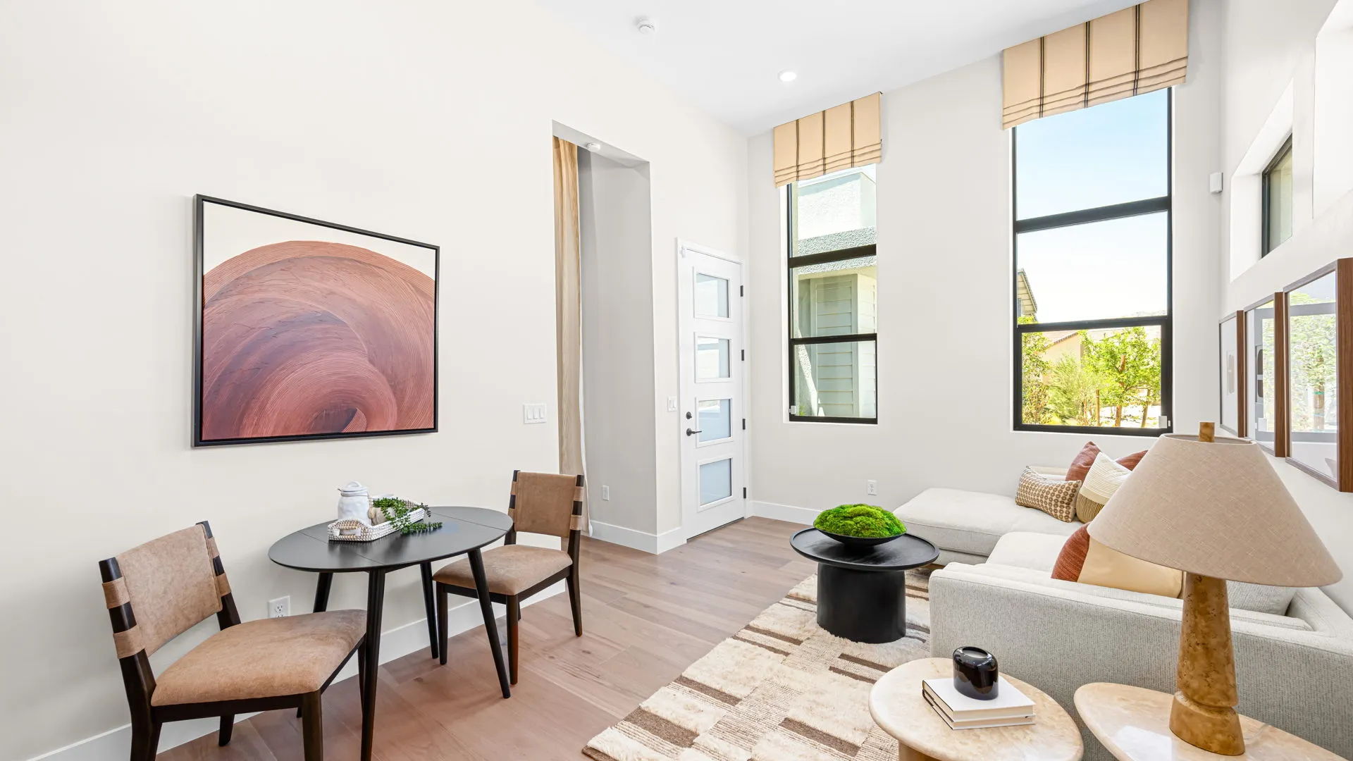 Suite of Monroe Model at Esplanade at Red Rock by Taylor Morrison (1) Bright living room with large windows, a white sofa, modern art, small dining table with two chairs, and light wood flooring. Natural light fills the space, creating an inviting atmosphere.