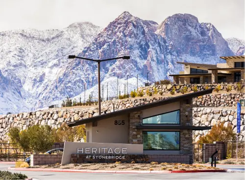 A modern stone building with a sign reading Heritage at Stonebridge stands in front of rugged, snow-capped mountains under a cloudy sky. Shrubs and a stone wall surround the area.