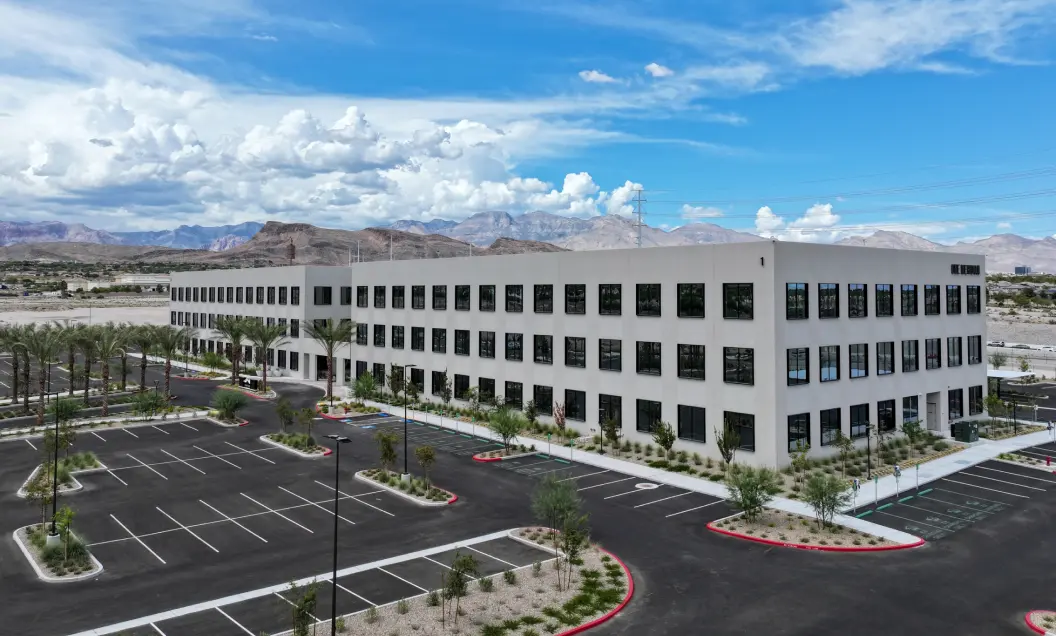 A large, modern three-story office building with many windows, surrounded by a mostly empty parking lot, palm trees, and desert landscaping, set against a backdrop of mountains and a partly cloudy sky.