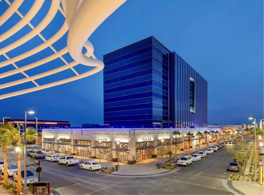 A modern shopping and dining complex at dusk features a large glass building in the background, an illuminated canopy overhead, palm trees, and parked cars lining the streets. The sky is deep blue.