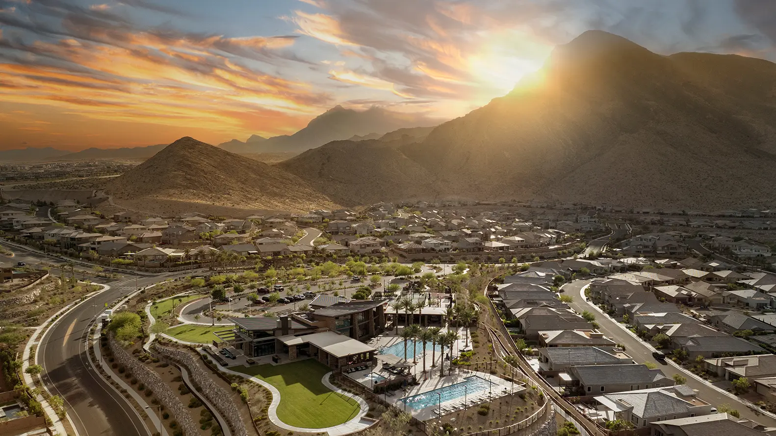 A suburban neighborhood nestles beside desert mountains at sunset, with houses, winding roads, green parks, and a swimming pool, all illuminated by the golden sunlight behind the peaks.
