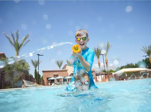 Two children playing in a swimming pool, with one child squirting water from a toy and both wearing swim goggles under a clear blue sky, palm trees and lounge chairs visible in the background.
