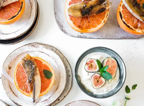 Several plates with broiled grapefruit halves topped with caramelized banana slices, and a bowl of yogurt garnished with fresh fig slices and basil, arranged on a white table.